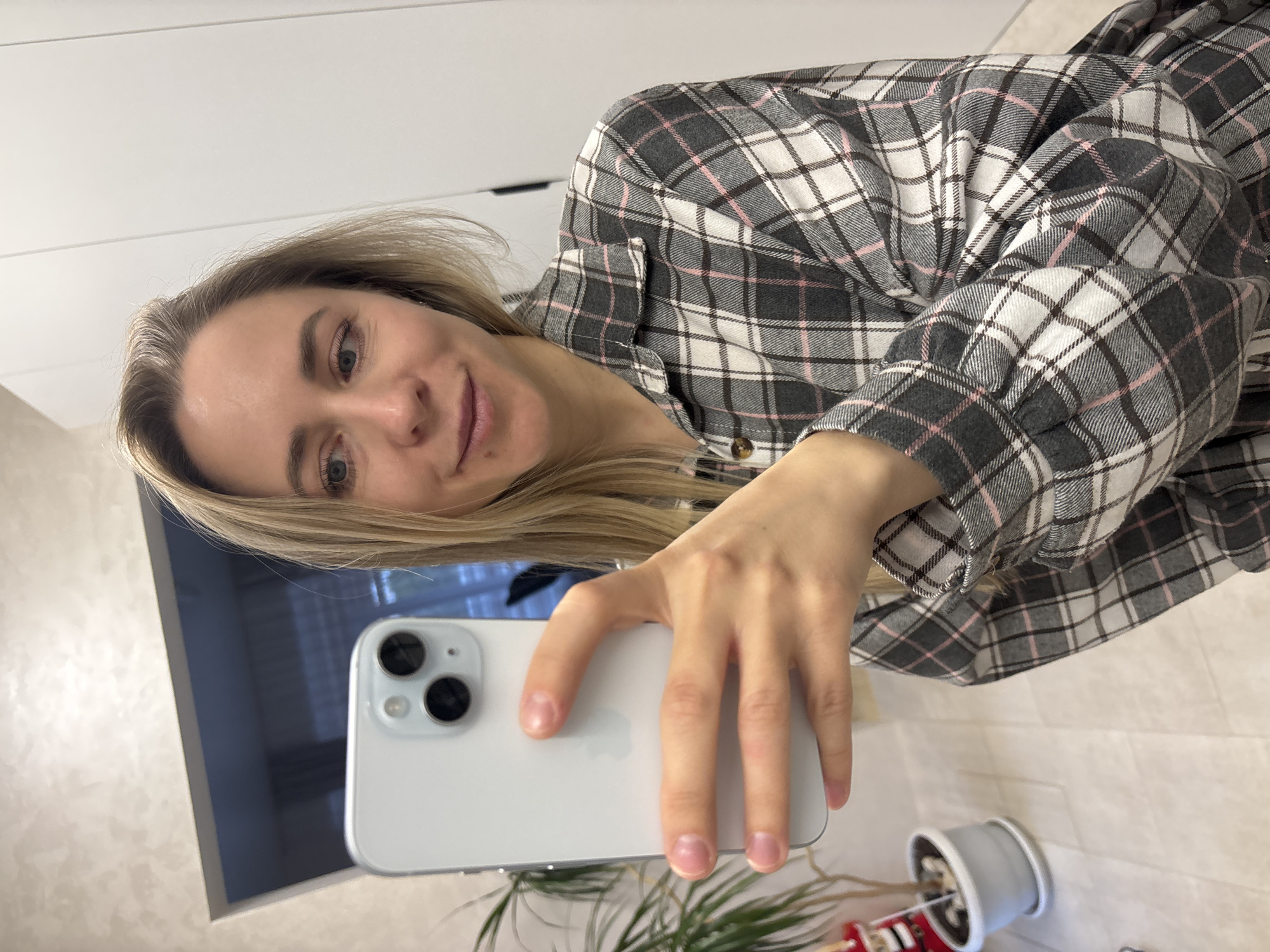 A woman with blonde hair is taking a selfie with a white smartphone. She is wearing a gray and white plaid shirt with pink accents. The background includes a white wall, a television, a plant in a gray pot, and a glimpse of the floor. The location is presumed to be in Chaikovsky, Russia.