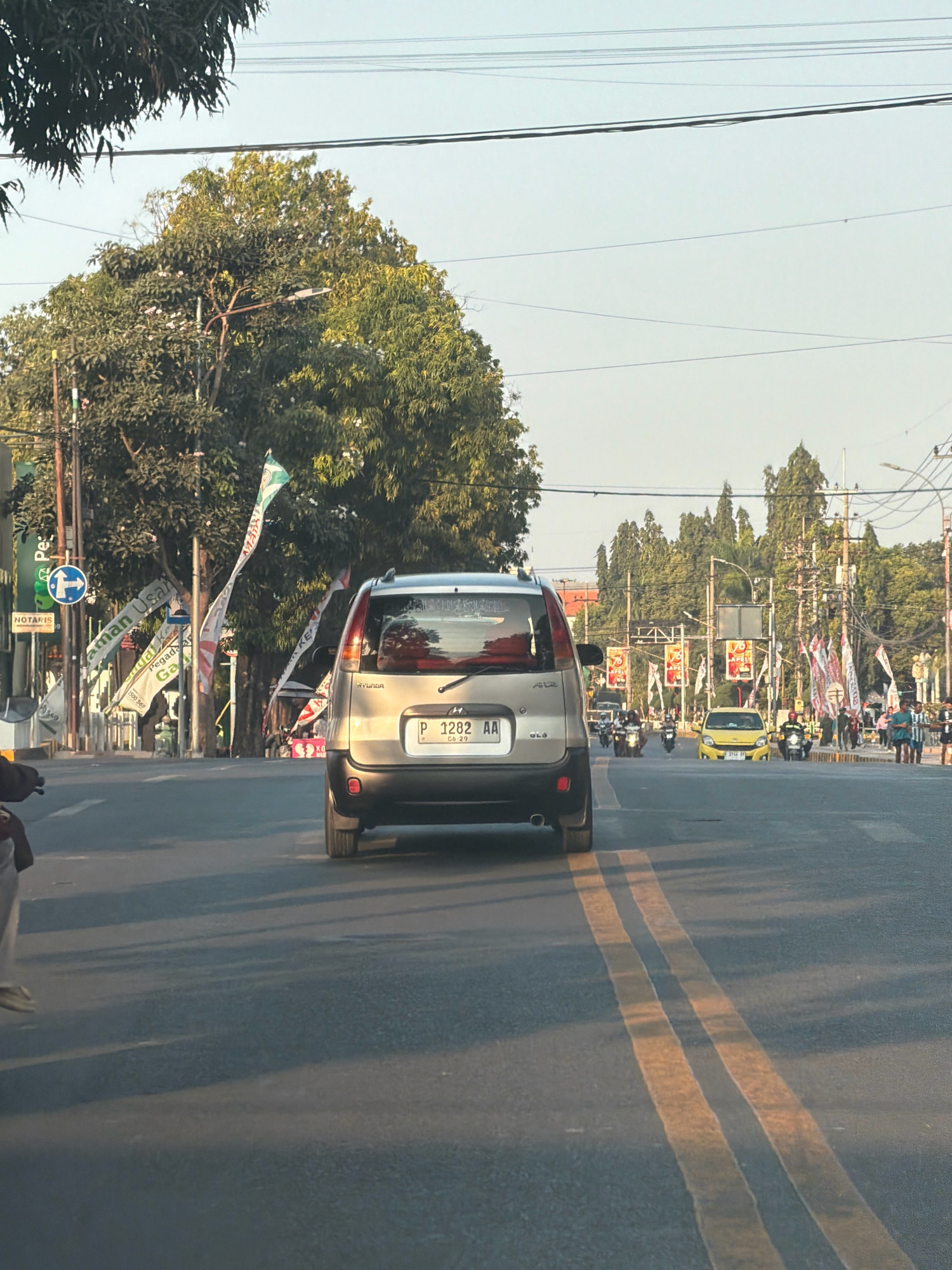 The image captures a street scene in Situbondo, Indonesia, during daylight hours. In the foreground, the rear of a silver Hyundai hatchback is visible, its license plate reading "P 1282 AA". The car is in motion on a paved road with double yellow lines. To the left of the car, a person wearing light-colored pants and shoes is partially visible, seated on what appears to be a motorcycle.
The roadside features several tall trees with dense green foliage. Banners and flags are prominently displayed along the street, some attached to poles and others fluttering in the breeze. One banner on the left displays text in Indonesian. A blue circular sign with a white arrow pointing left is also visible on a pole.
Further down the road, other vehicles are present, including several motorcycles and a yellow car. Pedestrians can be seen on the sidewalks and crossing the street. The background shows a line of trees and buildings, with overhead power lines crisscrossing the sky. The overall atmosphere suggests a busy urban environment.