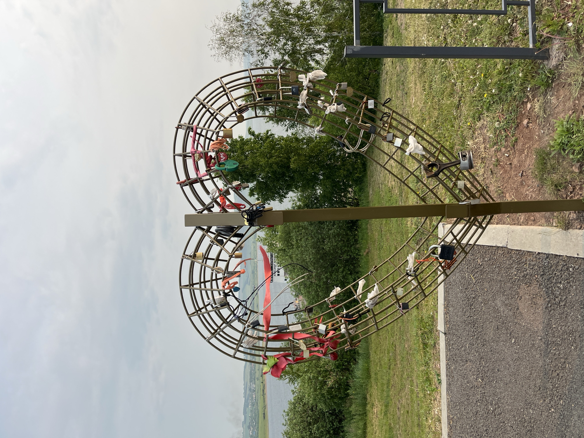A large, metal heart-shaped sculpture, adorned with numerous love locks and ribbons, stands outdoors in Novosyolovo, Russia. The sculpture is a central focus, positioned on a grassy slope overlooking a body of water, likely the Krasnoyarsk Reservoir, with a distant town visible on the opposite bank. The sculpture itself is constructed from curved metal bars forming the heart shape, with an internal grid structure where the locks and ribbons are attached. Several locks of various sizes and colors are visible, as are red, pink, and white ribbons tied around the metal framework. Some locks are weathered, suggesting they have been in place for some time. The environment is green and natural, with trees and grass surrounding the sculpture. The sky is overcast, indicating a cloudy day. The foreground shows a paved pathway and a metal railing. The overall scene conveys a sense of romantic tradition and public art within a scenic natural setting.