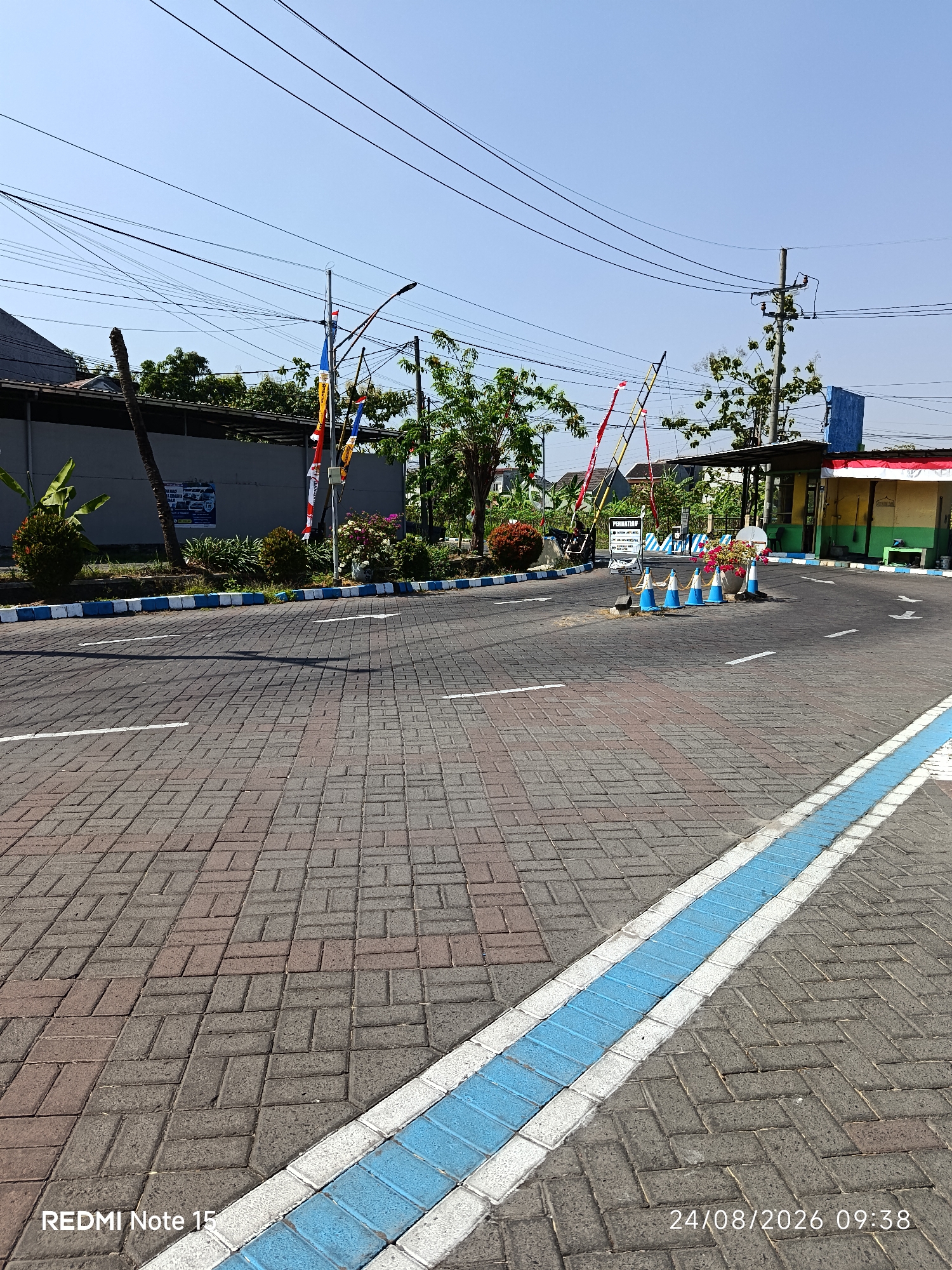 The image depicts a paved road junction in Keboananom, Indonesia, under a clear blue sky. The road surface is made of interlocking brick pavers, with a blue and white painted stripe along the right edge, guiding traffic. White arrows are painted on the road to indicate directional flow.
On the left side of the road, a grey wall is visible, partially obscured by lush green foliage and a palm tree. Several flags, including the Indonesian flag and other decorative banners, are flying from poles. Further along, there are trees and flowering bushes.
On the right side of the image, a small building with yellow and green walls serves as a guardhouse or entrance. A red and white awning extends from the building. Traffic cones, colored blue and white, are placed in front of the entrance, and a sign that reads "PERINGATAN" is visible. The scene is devoid of any visible people or vehicles, suggesting a quiet or inactive period. The overall environment appears to be a well-maintained outdoor area, possibly an entrance to a facility or a commercial establishment.