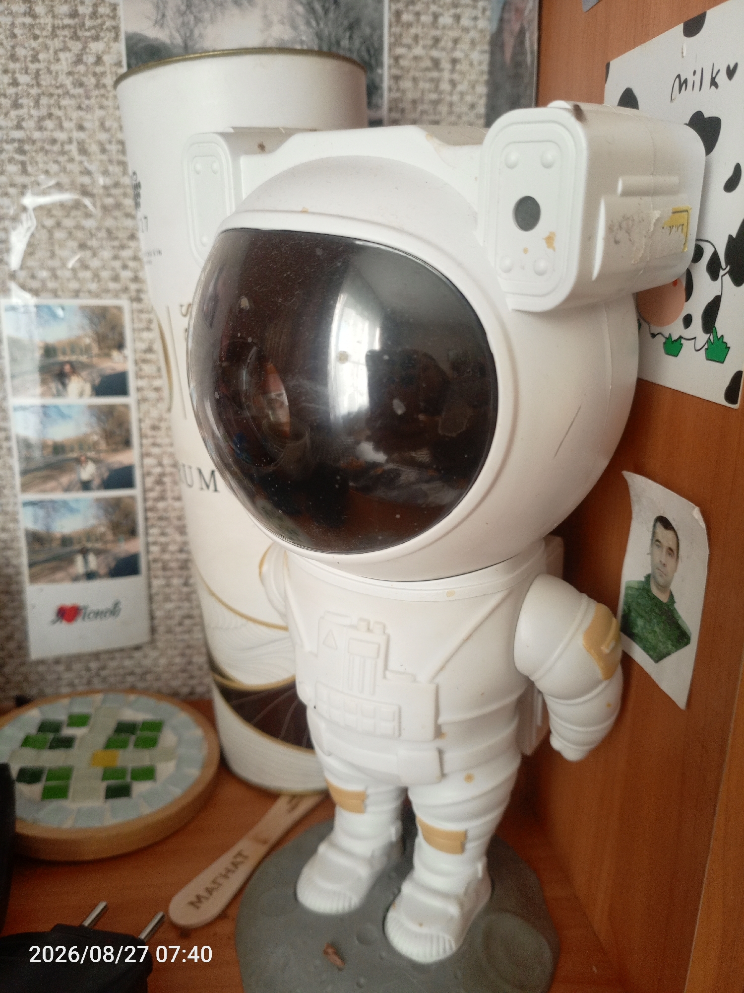 The image shows a toy astronaut figure standing on a grey, moon-like base. The astronaut is white with some yellow accents and a dark, reflective visor. To the left of the astronaut, there is a tall white cylindrical object with gold detailing and text that is partially obscured. Below and to the left of this cylinder is a strip of four small photos depicting people outdoors. A circular object with a mosaic of green and blue tiles is also visible on the left. A wooden object with the word "MATHAT" written on it lies horizontally in the foreground. In the background, to the right of the astronaut, is a wooden surface with a piece of paper that has a drawing of a cow and the word "Milk" written on it. Below this, a small printed photograph of a man in a military uniform is attached to the wooden surface. The timestamp "2026/08/27 07:40" is visible in the bottom left corner. The overall environment appears to be an indoor setting, possibly a shelf or desk, with wooden and textured surfaces. There is no explicit indication of the city of Pechory, Russia, within the image itself.