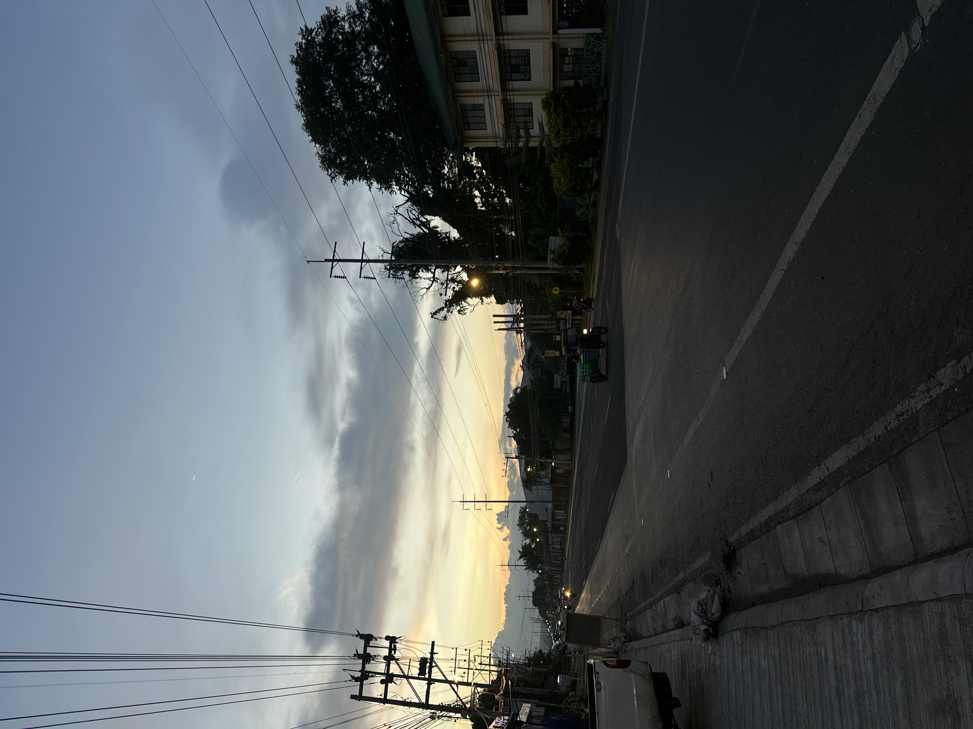 The image captures a street scene in Davao City, Philippines, during twilight. The sky displays a gradient of colors, from a pale yellow near the horizon to a dusky blue overhead, with scattered clouds. A crescent moon is visible in the upper portion of the sky. Utility poles with numerous wires run along the side of the road, dominating the left and central parts of the frame.
On the left side of the road, a white pickup truck is parked, and a gray concrete sidewalk with a curb is visible. Some discarded plastic bags and debris are present near the curb. The road itself is asphalt, with a visible white lane marker.
Several vehicles are present on the road, including a tricycle with passengers moving towards the viewer, and other cars and motorbikes further in the distance. Streetlights and building lights illuminate parts of the scene.
On the right side of the road, a two-story building with a light-colored facade, green roofing, and numerous windows is visible. Adjacent to it, there are trees and smaller buildings. The overall atmosphere suggests the end of the day in a semi-urban or suburban area of Davao City.
