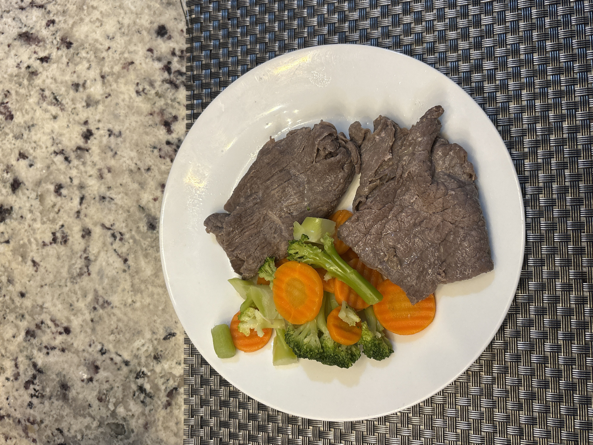 The image depicts a white plate containing cooked beef and sauteed vegetables (carrots, broccoli, and celery). The plate is positioned on a textured placemat. Part of the plate rests on a countertop with a mottled pattern. The location is not discernible.