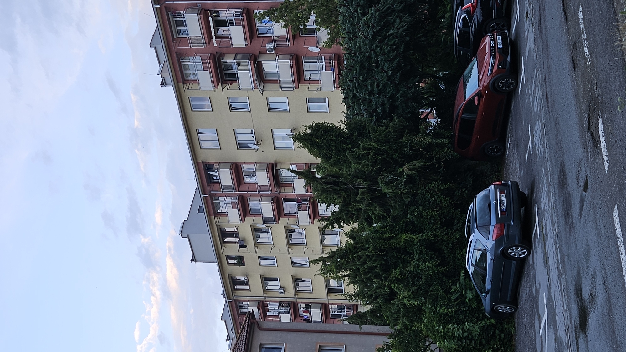 The image shows an apartment building in Prešov, Slovakia. The building is a multi-story structure with yellow and red facades, featuring multiple balconies with white railings. Several windows are visible, some with air conditioning units mounted outside. In front of the building, there is a row of dense green trees and shrubs. A parking lot is visible in the foreground, with several cars parked. One car is a dark gray station wagon, another is a reddish-orange SUV, and a third car, a black SUV, is partially visible on the right. The ground in the parking lot appears wet, possibly from recent rain, with painted parking lines visible. The sky is overcast with clouds.