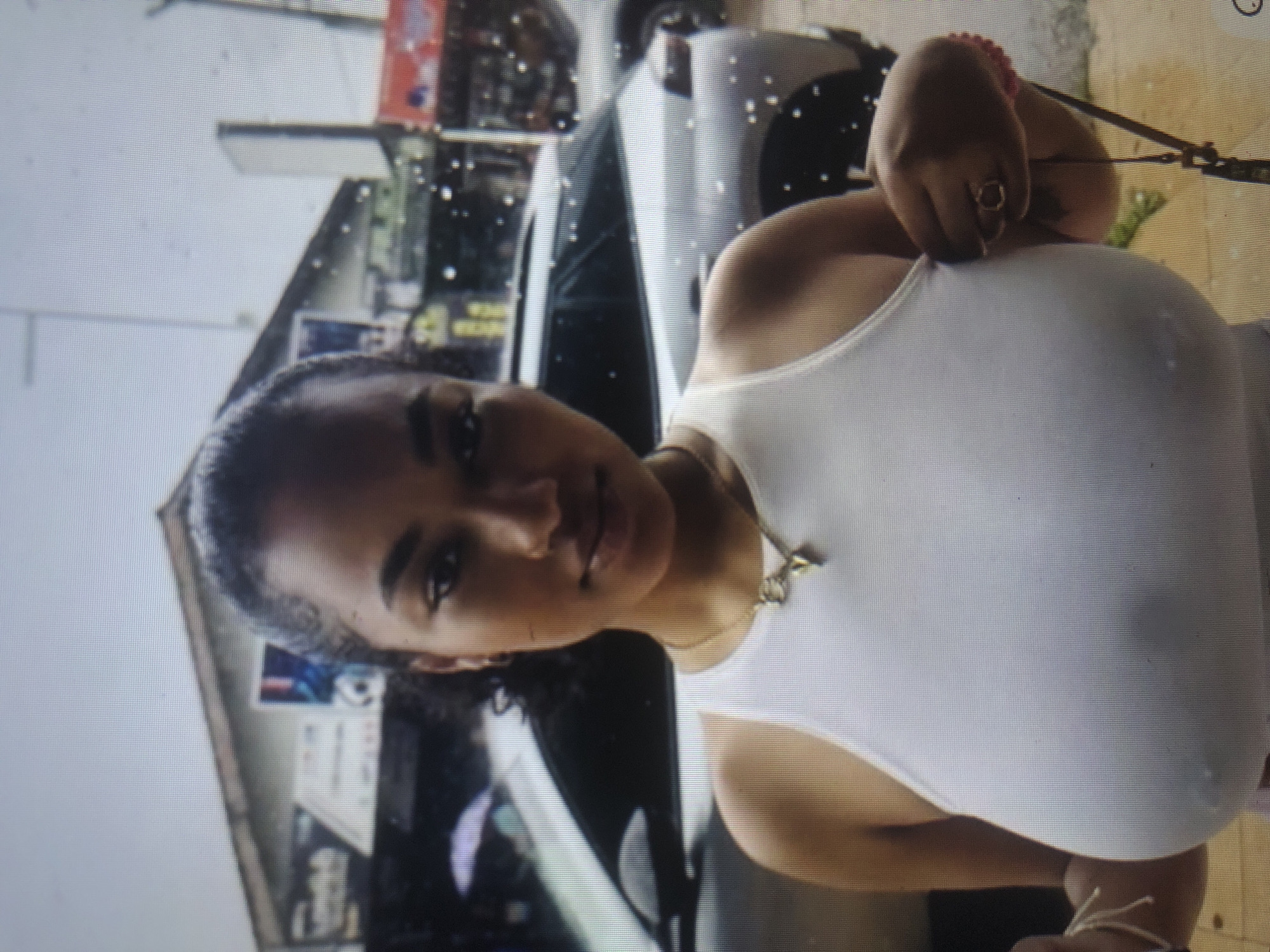 The media file shows a medium shot of a young Black woman from the chest up, angled to the right. She is wearing a white tank top and has curly dark hair. She is also wearing a gold necklace with a small pendant and a pink beaded bracelet on her right wrist, with a ring on her right index finger. Her expression is neutral, and she appears to be outdoors, possibly in an urban environment, with buildings and a car visible in the background. The lighting suggests it might be daytime. The background contains blurry details that could be signs or posters on the buildings. There is no discernible text in the image. The setting appears to be a street in Barakin Lerawi, Nigeria.