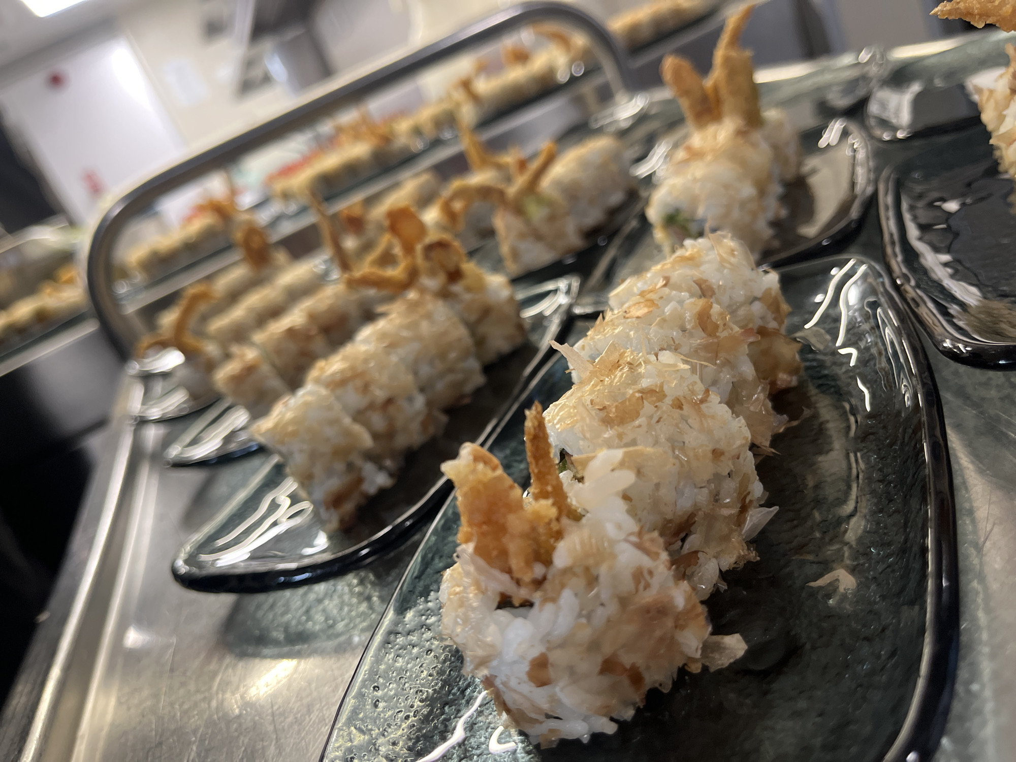 The image presents an angled, close-up view of several rows of sushi rolls arranged on dark, textured rectangular plates. The sushi in the foreground is sharply focused, showcasing what appear to be Ebi Tempura rolls, identifiable by the distinct fried shrimp tails protruding from the top of the rolls. The rolls are generously coated with a light, flaky topping, possibly toasted coconut flakes or crispy breadcrumbs, which adds texture to the white rice.
The plates are displayed on a reflective, metallic surface, likely a commercial food counter or buffet setup. In the mid-ground, another set of similar sushi rolls is visible on a parallel display, slightly blurred. The background remains out of focus, revealing indistinct light-colored walls and surfaces, suggesting an indoor commercial kitchen or a serving area. The lighting is artificial, creating soft reflections on the metallic counter and the sleek plates. No people are visible in the scene, and there is no discernible text. The overall setting implies a food service environment in Sofia, Bulgaria, where these sushi rolls are prepared for display and consumption.