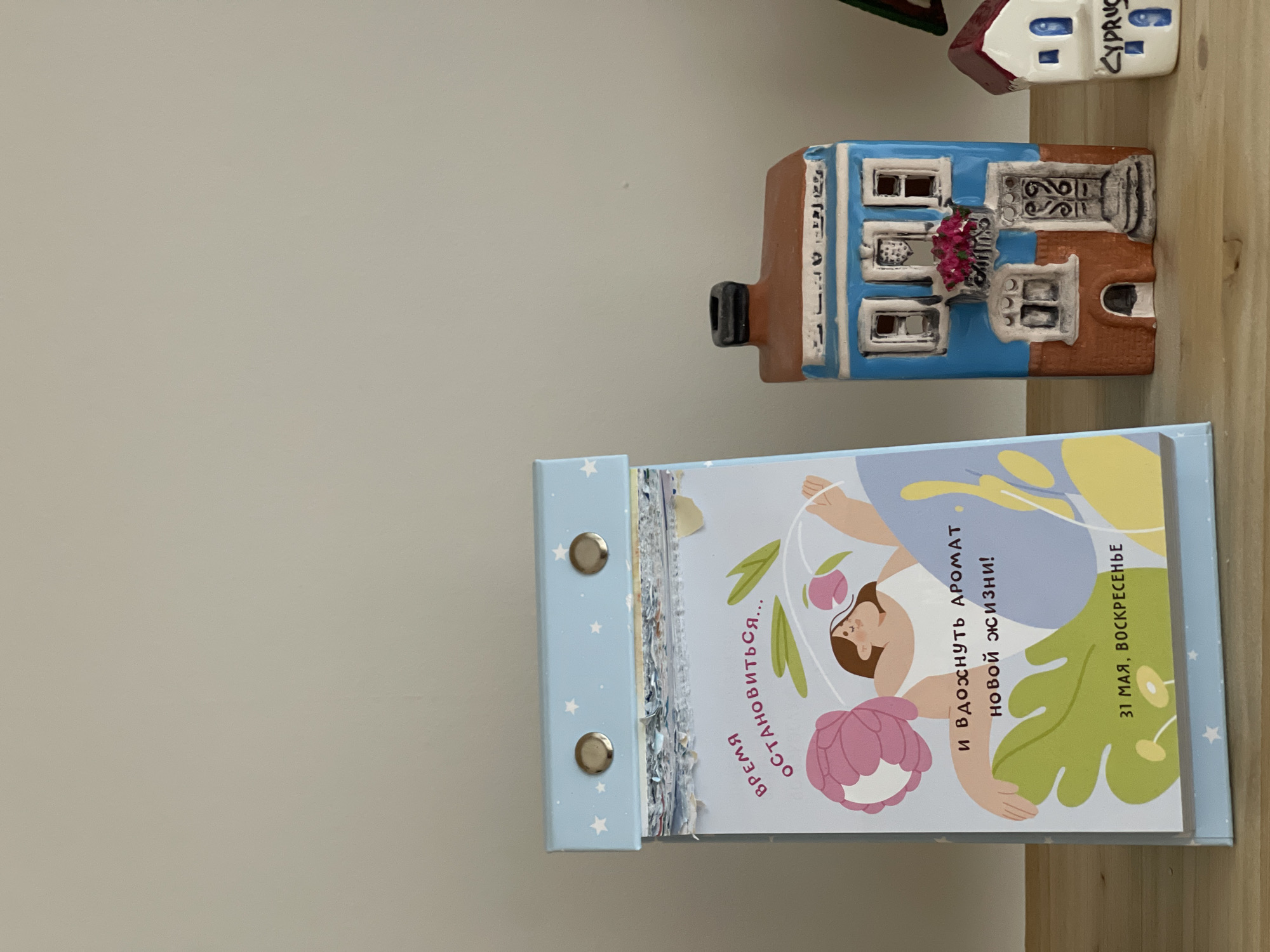 A still life of tourist souvenirs and a desk calendar on a light wooden surface, set against a pale gray wall. The items are arranged horizontally, with the camera positioned at a slightly above eye level.
The desk calendar is the largest object in the frame, light blue with small white star patterns on its edges. Its open page displays a stylized illustration featuring a person lying amidst botanical elements, and contains Cyrillic text in dark lettering. The visible text includes: "Время остановиться..." (Time to stop...), "и вдохнуть аромат новой жизни!" (and inhale the aroma of a new life!), and "31 мая, воскресенье" (May 31, Sunday).
Above the calendar are two small ceramic house figurines, typical tourist souvenirs. The house closest to the camera has a brown tiled roof and a blue facade with painted windows and decorative elements. The house further back has a white facade with a brown roof and the word "CYPRUS" inscribed.
The scene is well-lit, and the colors are clear and muted. The setup appears deliberate, as if for a casual snapshot or a product display.