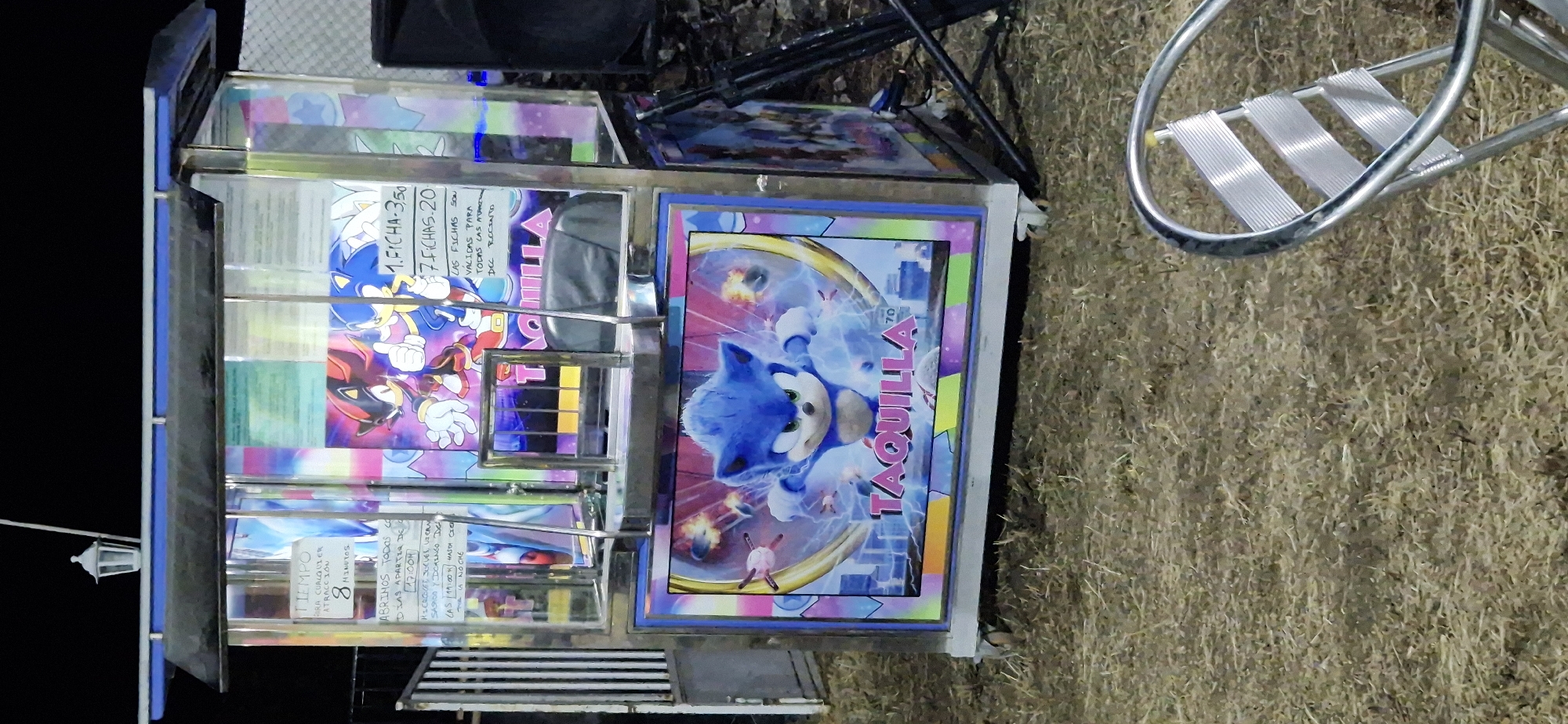 The image shows a ticket booth decorated with Sonic the Hedgehog characters at a fair or amusement park. The booth has glass panels displaying pricing information for rides or attractions. On the front of the booth, there is a large graphic of Sonic in an action pose. To the right of the booth, a dark speaker or sound system is visible, and a metal chair with a slatted seat is in the foreground. The ground is covered with dry grass or straw. The overall setting appears to be outdoors at night, possibly during an event or festival. The text on the booth suggests it's a ticket sale point ("TAQUILLA" means "ticket office"). The text also indicates operating hours and prices, with options for one ticket for 3.50 and seven tickets for 20. It also specifies that tickets are valid for all attractions. The image is undated.