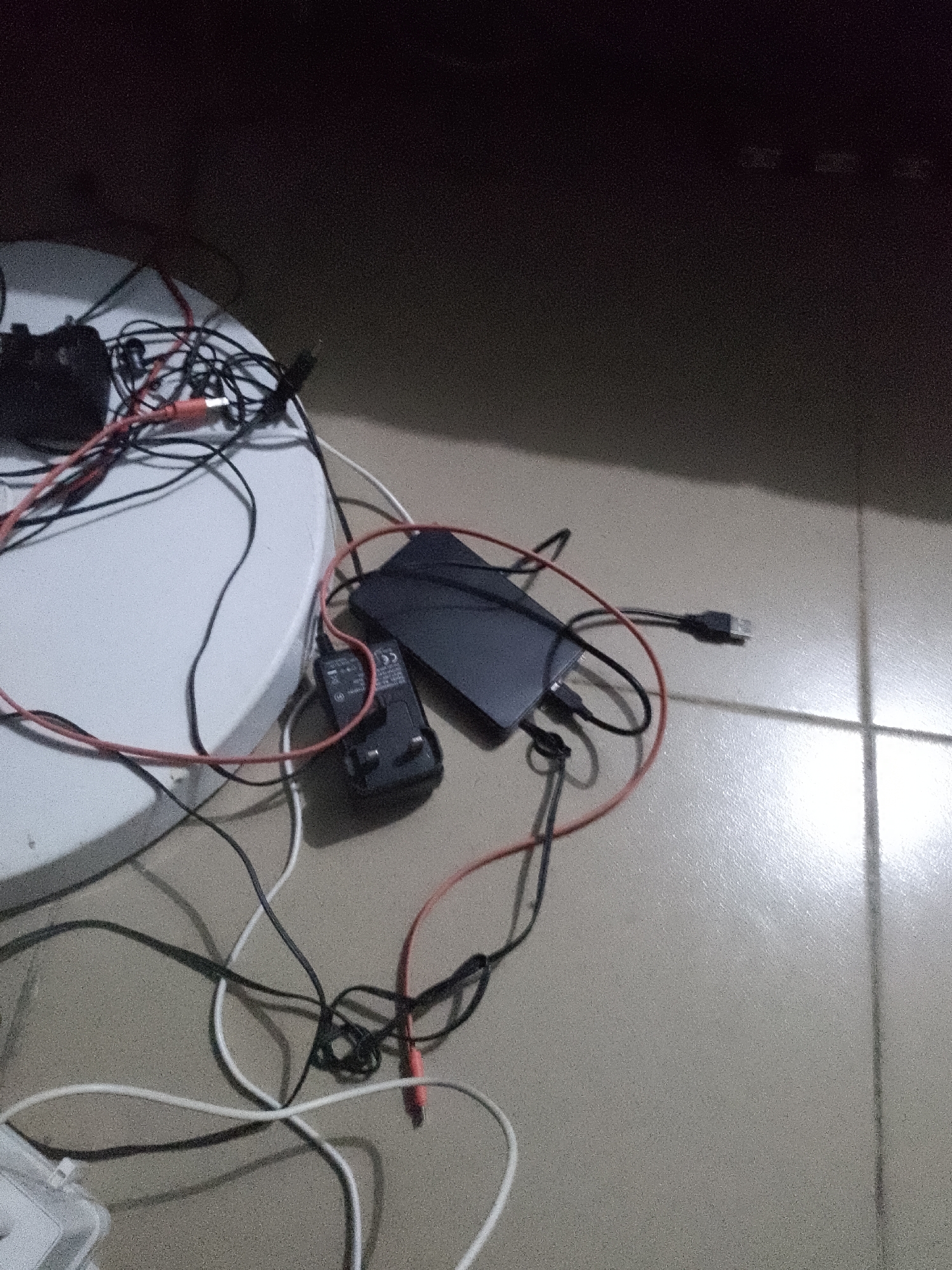 An overhead, dimly lit, high-angle medium shot captures a chaotic collection of electronic wires and devices scattered across a white circular surface and a tiled floor. A black power brick with a CE marking and prongs for an electrical outlet is visible, connected to a black cable. A dark rectangular electronic device, possibly a portable hard drive or a similar storage device, is also present, with a black USB cable extending from it. Red and black wires snake across the scene, tangled with white cables. The lighting suggests it is nighttime, with some light reflecting off the tiled floor. The context of Jauro Yinu, Nigeria, is inferred from external metadata and not visually evident within the image itself. There are no people visible in the frame, and no discernible actions or interactions are taking place. The environment appears to be an indoor setting, possibly a room or a study area.