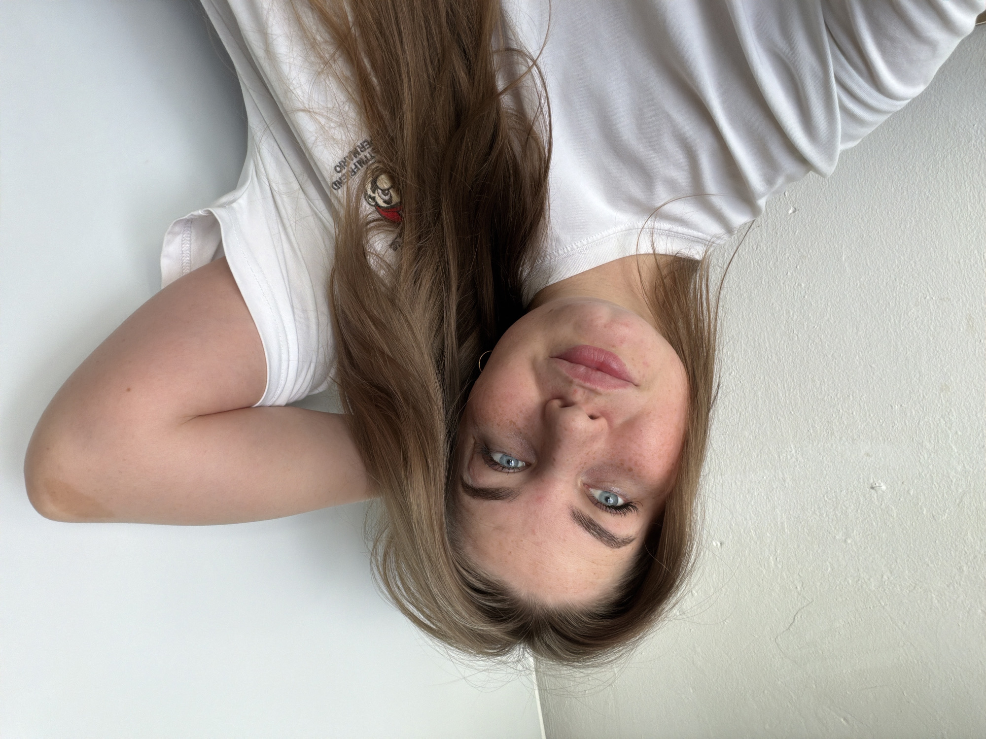 A young Caucasian woman with long, light brown hair and blue eyes is looking into the camera. She has fair skin with freckles, rosy cheeks, and is wearing a white t-shirt. Her right arm is bent behind her head. She appears to be indoors, with a textured white wall behind her. There is no indication of the city of Chaikovsky, Russia, or any specific urban environment.
