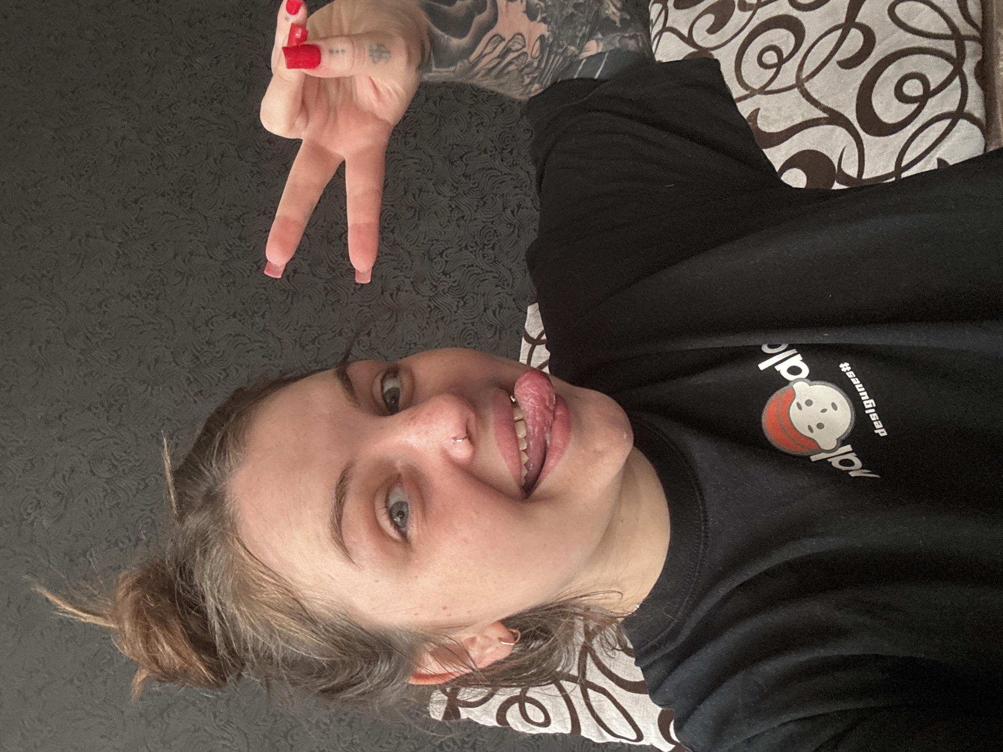 A woman with a nose ring, a bun hairstyle, and red manicured nails is sticking out her tongue while making a "peace" or "V" sign with her fingers. She is wearing a black long-sleeved shirt with a design on it, lying on a patterned surface. There is also a tattooed arm visible. The location, Chornomorskoe, Ukraine, cannot be determined from the provided image.