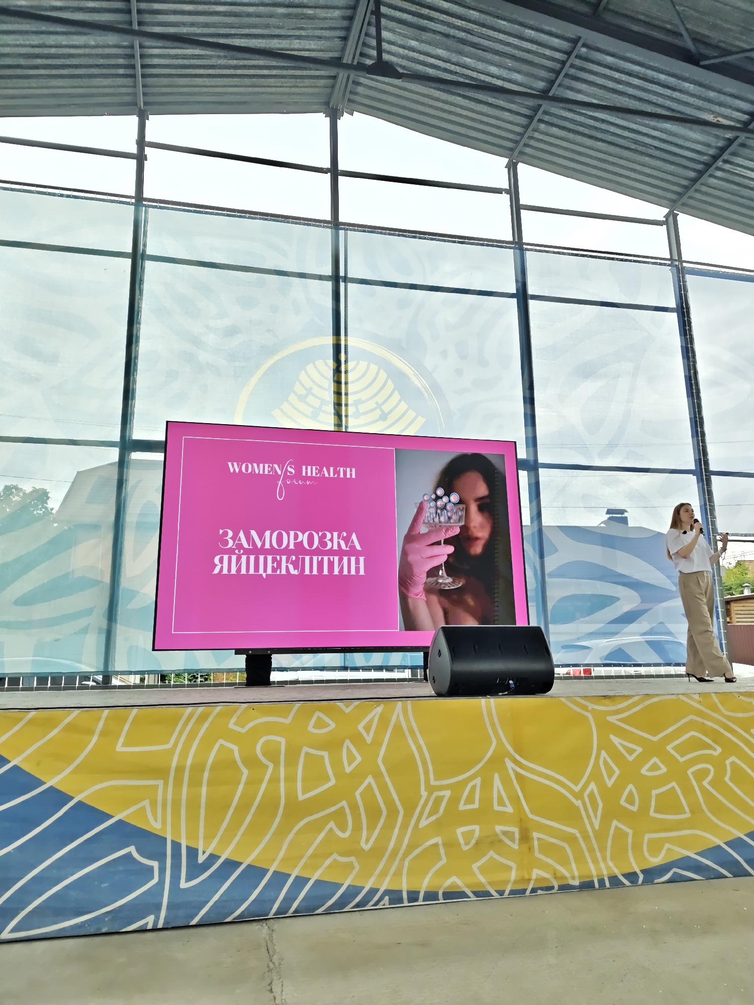 The image captures an outdoor or semi-outdoor event taking place on a stage in Vinnytsia, Ukraine. A female speaker is presenting next to a large LED screen, which displays information about "WOMEN'S HEALTH forum" and "EGG FREEZING" in Ukrainian.
The scene is set on a raised stage with a facade adorned with an abstract pattern of white outlines over a diagonally split yellow and blue background. A large, rectangular LED screen stands centrally on the stage, displaying content with a prominent pink background. The left portion of the screen features white text: "WOMEN'S HEALTH forum" at the top, and "ЗАМОРОЗКА ЯЙЦЕКЛІТИН" (Ukrainian for "EGG FREEZING") below. The right portion of the screen shows a close-up image of a woman with long dark hair, wearing pink gloves, and holding a clear glass containing numerous small, light-pink spherical objects.
To the right of the screen, a woman with straight, light-colored hair, wearing a white short-sleeved top, light beige wide-leg trousers, and dark high-heeled shoes, stands and addresses an unseen audience. She holds a dark microphone in her left hand and gestures with her right. A black stage monitor speaker is positioned on the stage floor in front of the LED screen, to the speaker's left.
The background consists of large, multi-paned glass walls, through which an overcast sky and distant green trees and building structures are visible. Reflections on the glass include a large, circular gold-colored emblem or logo and various abstract blue and white patterns. The ceiling above the stage is a corrugated metal structure supported by exposed metal beams. The overall lighting suggests natural daylight under an overcast sky.