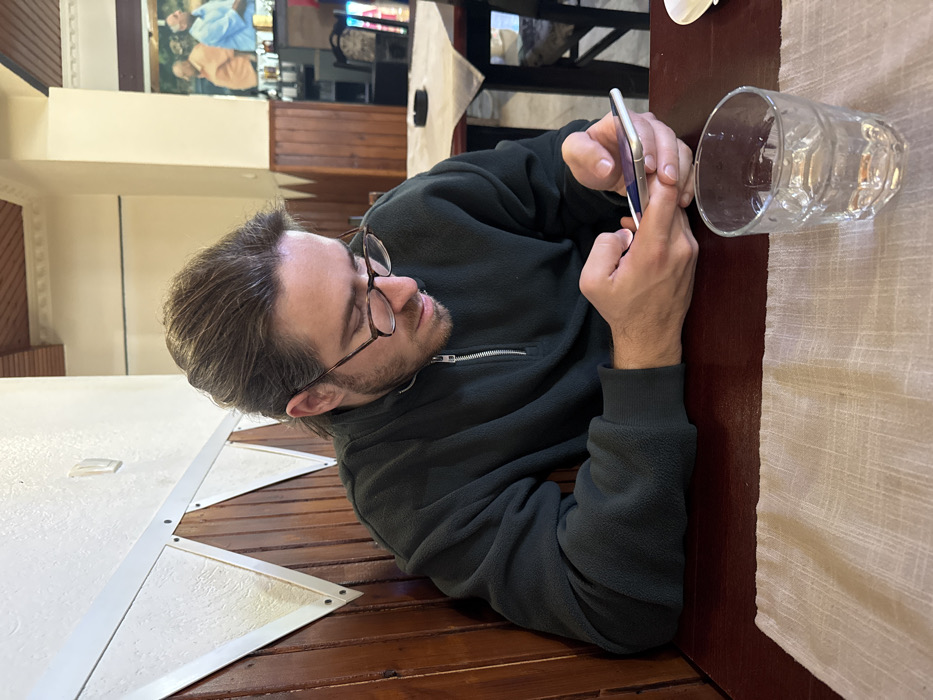 A male individual is seated at a dark brown wooden table within an indoor establishment in Tutin Municipality, Serbia. He has medium-length, light brown hair, wears dark-rimmed eyeglasses, and is dressed in a dark green fleece quarter-zip pullover. His focus is directed downwards as he operates a white smartphone with both hands. A clear, faceted glass containing water rests on a light beige woven placemat to his right. The immediate background features a wall with vertical dark wood paneling topped by a white border and decorative white triangular relief panels. The deeper background reveals additional dining tables with light-colored tablecloths and dark chairs, and a wall mural depicting two men is visible further into the room, beneath a wooden ceiling.