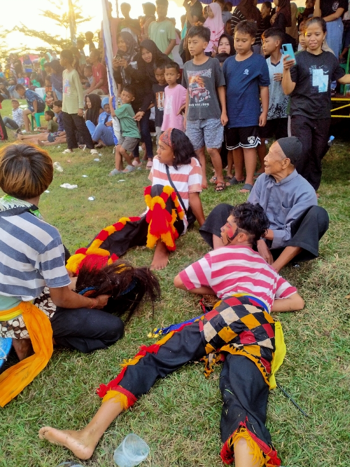 This image captures an outdoor scene in Glonggong, Indonesia, on a grassy field, likely during a cultural performance or event. The foreground features four main individuals on the grass. On the left, an individual with short, light brown hair, wearing a white and blue striped shirt, kneels facing away from the viewer while handling a dark, shaggy object, possibly a costume prop. Next to them, a performer with long dark hair and a patterned headband, dressed in a white and orange striped shirt and black pants adorned with red and yellow fringed fabric, sits barefoot and looks to the left. In front of them, another performer lies prone, propped on an elbow, facing left. This individual has short dark hair, face paint with white and red marks, wears a red and white striped shirt, and black pants with a prominent multi-colored checkered pattern (black, white, red, yellow) on the lower body, also with red and yellow fringe at the ankles and barefoot. Adjacent to this prone performer, an elderly man in a dark cap, a light grey long-sleeve shirt, and dark pants sits cross-legged, observing the scene. The grass contains minor debris, including a clear plastic cup. In the midground and background, a crowd of approximately 30-40 individuals of various ages, including children and adults, stand and sit, spectating. Many women in the crowd wear hijabs. On the far right of the crowd, a woman holds a blue smartphone, appearing to record or photograph the event. The scene is illuminated by natural light, suggesting late afternoon.