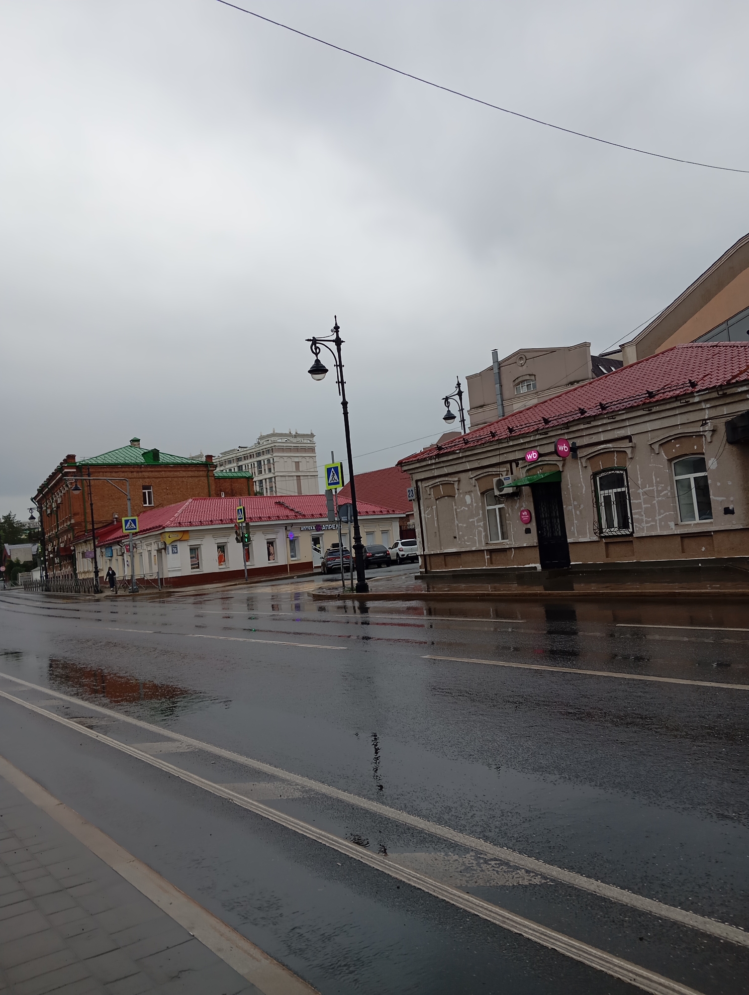 The image depicts a street scene in Orenburg, Russia, on a cloudy, overcast day, suggesting recent or ongoing rain, as evidenced by the wet asphalt and reflections. In the foreground, a wet road with white lane markings takes up a significant portion of the frame. To the right, a single-story building with a red tiled roof and a light-colored facade is visible, featuring several windows and an entrance. Two purple circular logos with "wb" are displayed on the facade. Further down the street to the left, another building with a red tiled roof and a white facade is seen, with a sign that reads "АПТЕКА АПРЕЛЬ" (Pharmacy April). Adjacent to this is a taller, older brick building with a green roof. A pedestrian, wearing dark clothing, walks on the sidewalk near the brick building. Traffic lights and pedestrian crossing signs are present. In the background, taller buildings are visible, including a modern multi-story structure. A black lamppost with an ornate design stands on the right side of the street. A thin wire stretches across the top of the frame. The overall atmosphere is somber and wet, typical of a damp urban environment.