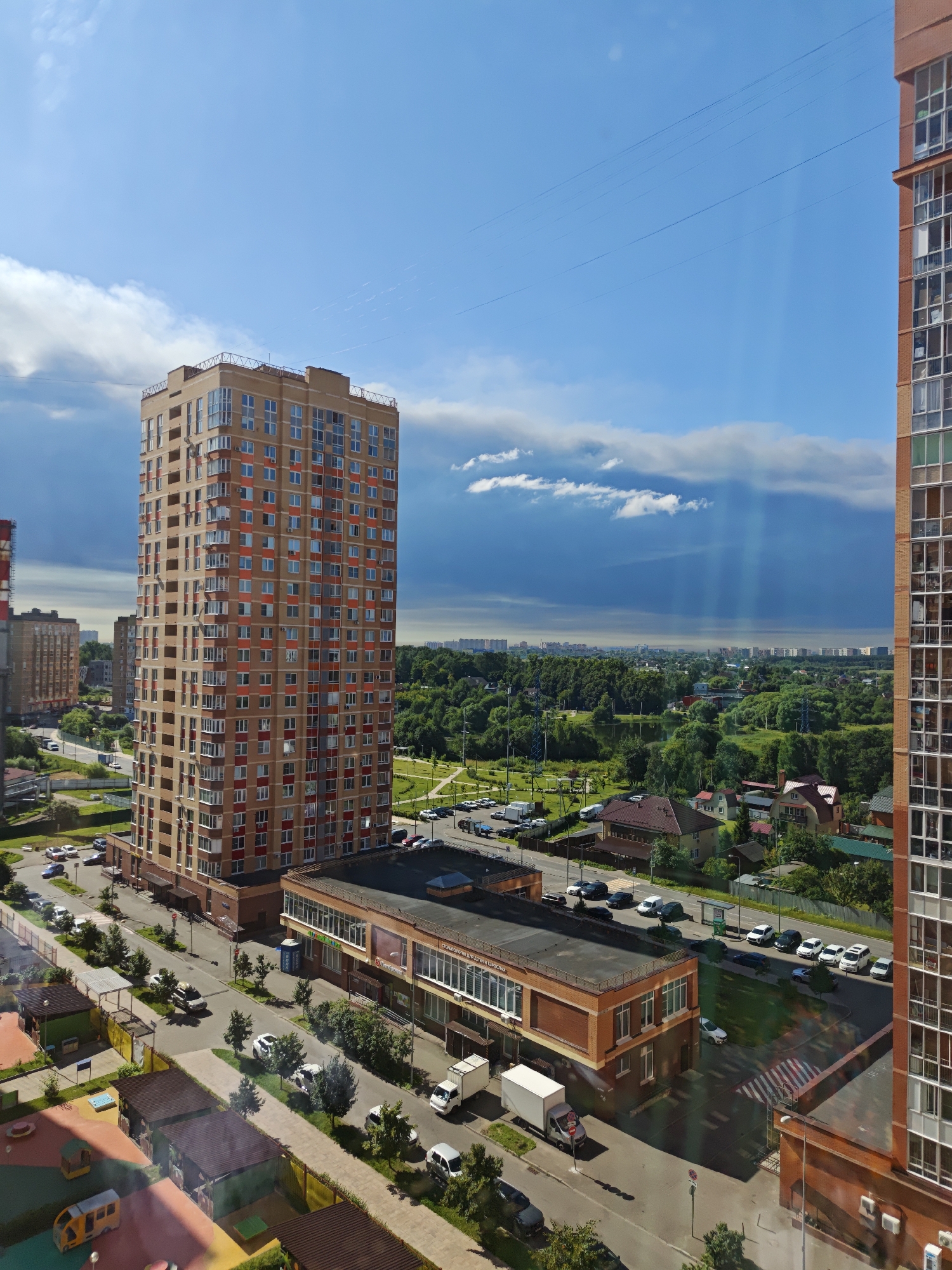 The aerial view captures a residential and commercial area in Shcherbinka, Russia, under a partly cloudy sky with visible sunbeams. Dominating the foreground is a tall, multi-story residential building with a light brown facade and red accents around the windows. To its left, a slightly shorter residential building is also visible, and a taller, partially completed building with a reddish-brown brick exterior stands on the far right.
In the middle ground, a low-rise commercial building with a brick facade and large windows runs horizontally across the frame. Several cars are parked in front of it. A street runs alongside this building, with more parked cars and some trees. Further into the distance, a dense green forest stretches across the landscape, punctuated by scattered houses and the faint outlines of more distant urban structures.
The lower part of the image shows a section of a residential complex with children's play areas featuring colorful surfaces and small structures. A yellow bus-like toy is visible on one of these areas. A sidewalk or path borders these play areas, with more trees and parked cars along the street. The scene appears to be from a summer day, with clear weather and lush vegetation.