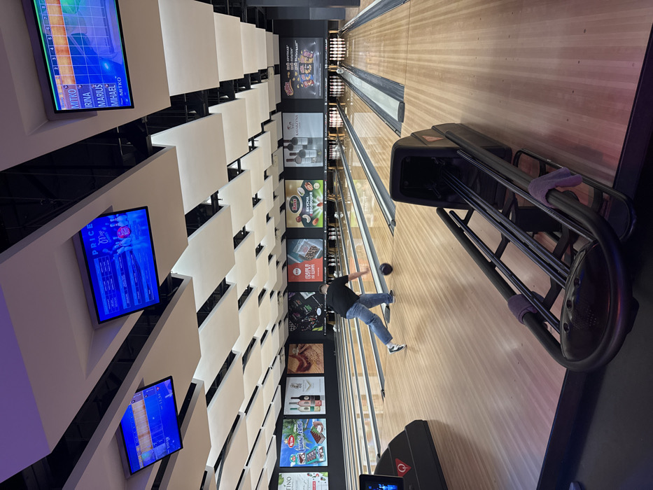 The scene depicts the interior of a modern bowling alley located in Sofia, Bulgaria. Multiple polished wooden bowling lanes extend into the background, terminating at pin setups. The ceiling features a complex arrangement of angled, white rectangular panels and integrated lighting.
In the mid-ground, a male individual is captured in a dynamic pose, releasing a dark bowling ball onto the lane. He wears a black short-sleeved shirt, blue jeans, and white bowling shoes. His body is bent forward, with his right arm extended towards the lane and his left leg extended behind him. The bowling ball is visible rolling on the lane immediately to his right.
In the foreground, on the right, a black bowling ball return mechanism is visible, featuring parallel metal rails. Two purple towels are draped over these rails.
Three large, rectangular monitors are suspended from the ceiling. The central screen displays "PRICE" at the top with what appear to be pricing tables and a graphic of bowling pins and a ball. The screen on the far right displays a bowling score interface with names "MITKO," "IRINA," "MARIUS," and "MIHAEL."
Along the dark wall above the lanes, a series of large, brightly colored advertisement posters are displayed. These feature various Bulgarian brands and products, including "Big Bon" chocolate, "Katarzyna Estate" wine, "Rois" nuts with "ВКУСНА ЕВОЛЮЦИЯ" (Tasty Evolution), and "Крушата" snacks. The environment is well-lit and appears clean and well-maintained. No other individuals or direct interactions are visible.