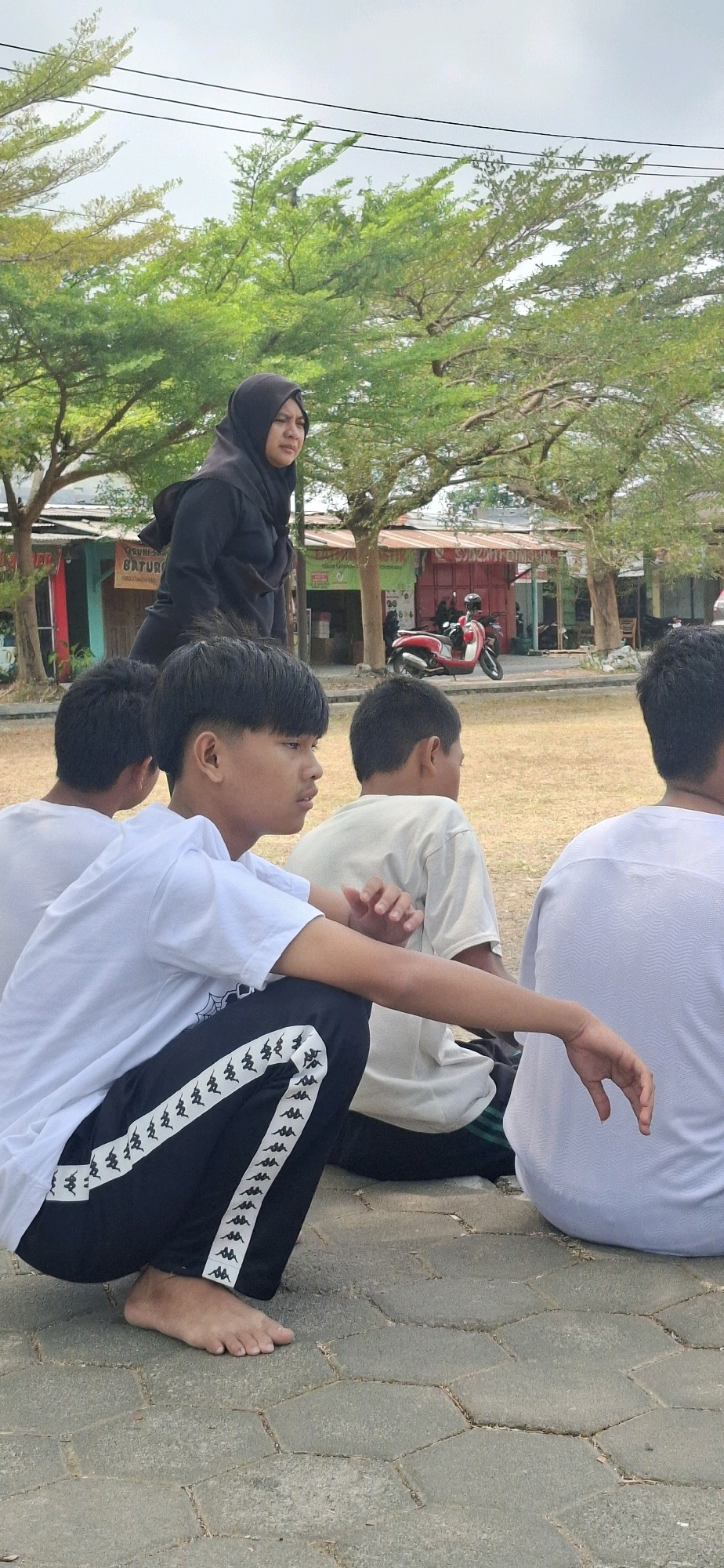 In Baturan, Indonesia, under a cloudy sky, four young males are seated on a paved area with a grassy field behind them. The foremost individual is squatting, facing right, wearing a white t-shirt and black pants with white stripes. His left hand rests on his right knee. To his left, another male is also seated, facing right. The third male, also seated and facing right, is partially visible behind the first. The fourth male is seated on the far right, facing away from the camera.
In the background, a woman wearing a black hijab and black clothing stands. She is looking towards the left. Behind her, several low-rise buildings with corrugated roofs are visible, along with trees. A red and white motorcycle is parked in front of one of the buildings. Power lines are visible crisscrossing the sky. The overall environment appears to be an outdoor public space, possibly near shops or a community area.