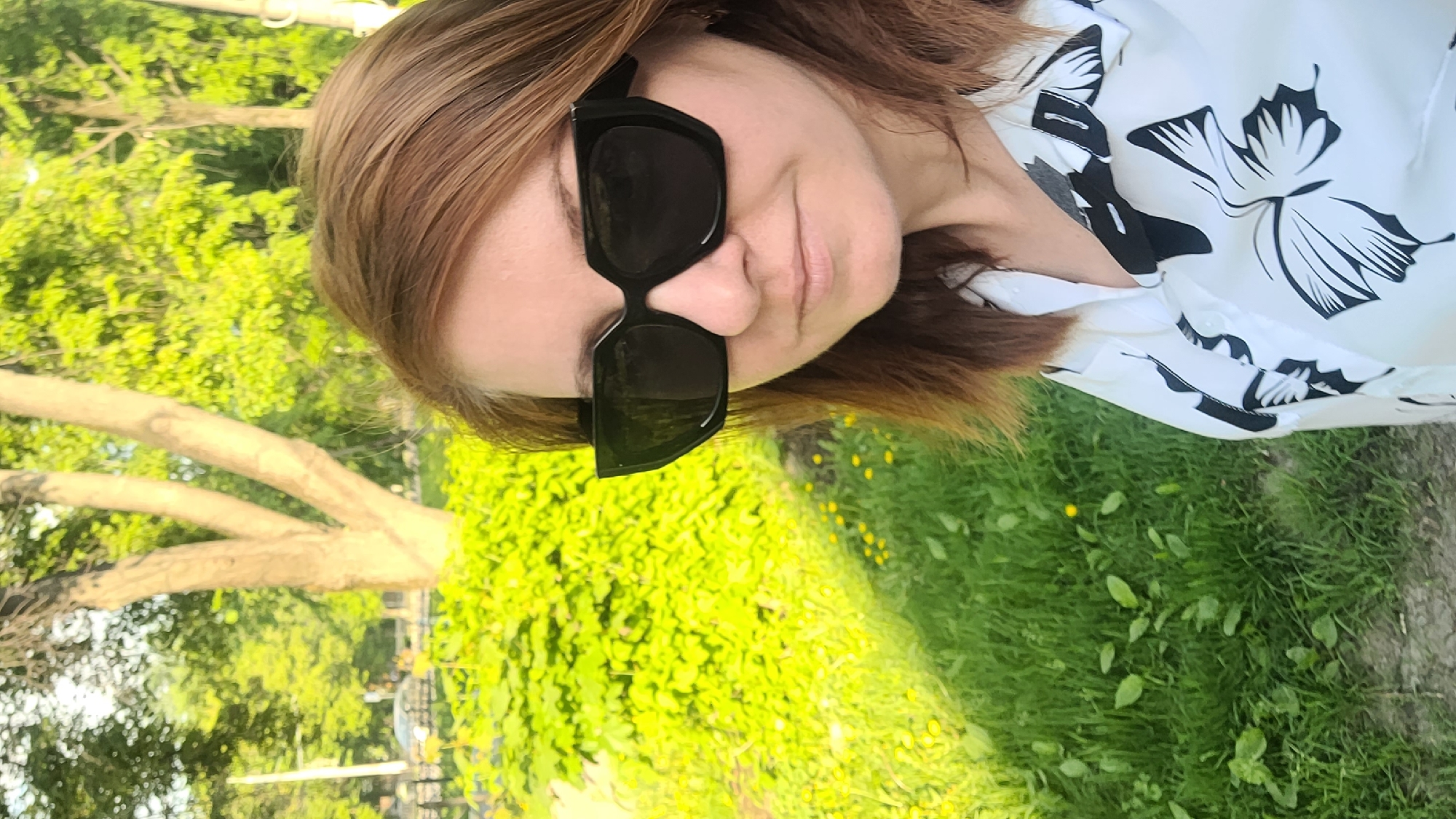 A woman wearing sunglasses and a white shirt with black floral patterns stands outdoors on a grassy area. Behind her is a green space with trees, possibly a park or similar green area. The location is the city of Unknown, Unknown.