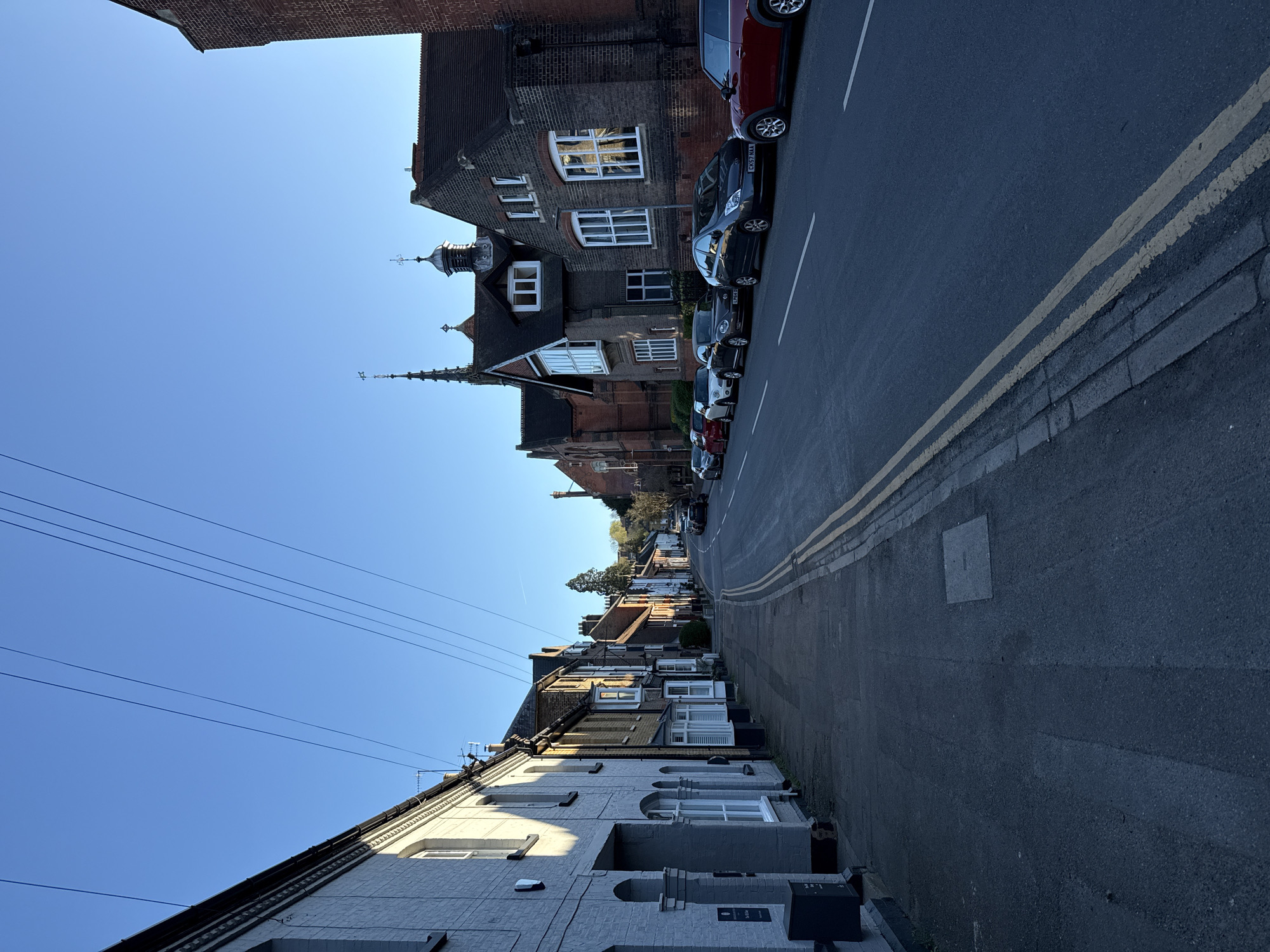 The image shows a street scene in Dacorum, United Kingdom. The street is lined with houses and parked cars. A double yellow line runs along the side of the road. Above, power lines are visible. The sky is clear and blue. The buildings vary in architectural style, including both brick and painted facades. A spire is visible in the distance. The road surface appears to be asphalt. The day is sunny.