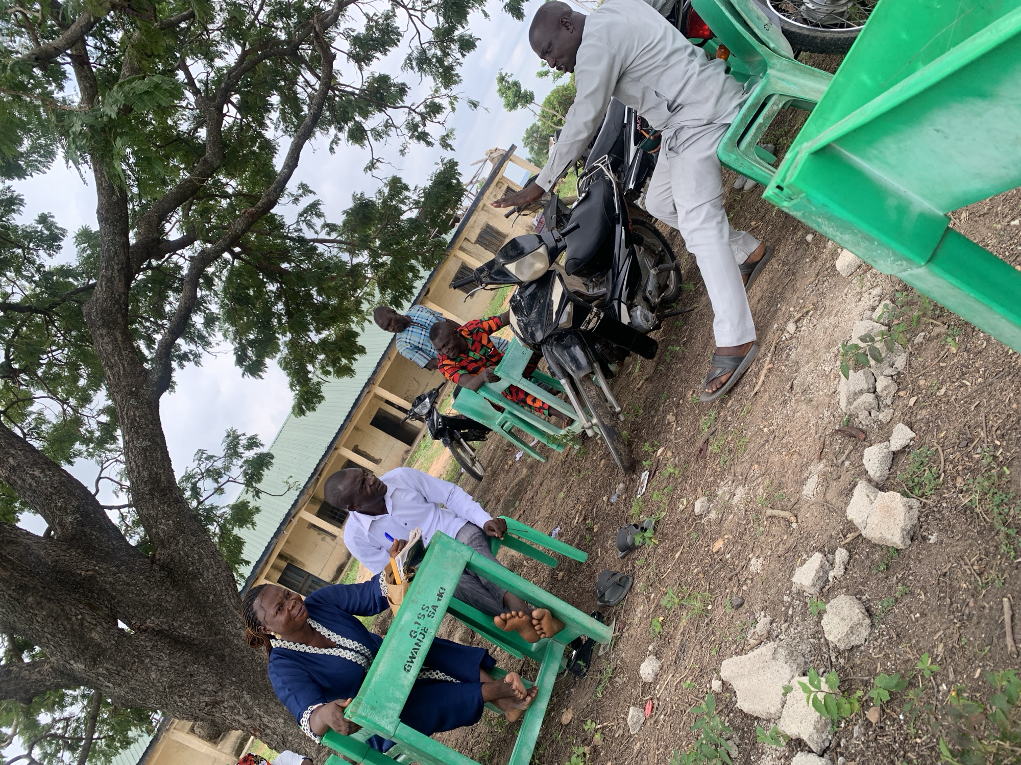 In Akwanga, Nigeria, under the sprawling branches of a large tree, four men and one woman are gathered outdoors near a light-colored building with a green roof. Two of the men are seated at bright green plastic tables, one writing in a notebook, the other conversing. A motorcycle is parked to the right. The ground is covered with dirt, small rocks, and sparse vegetation. One man is seated on a green plastic chair next to a motorcycle, gesturing with his hand, while another man stands by the motorcycle, appearing to speak to him. A fifth man stands near a table on the left side of the image, wearing a checkered shirt. The woman is seated at a table on the left, wearing a blue dress.