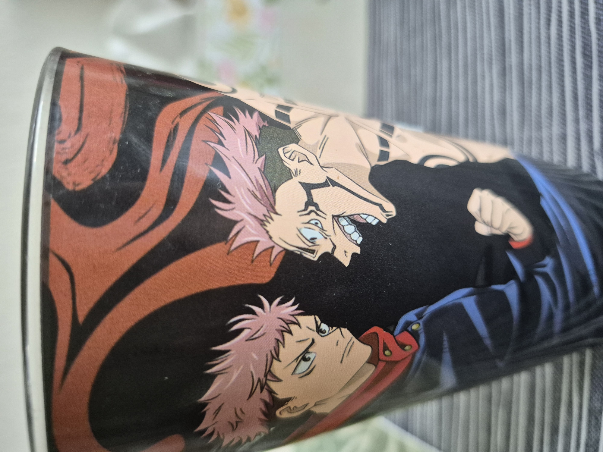 The media file shows a close-up, angled view of a cylindrical object, likely a container or a can, adorned with an anime-style illustration. The artwork features two characters with spiky pink hair. One character, positioned higher and in the background, has an aggressive expression with wide, shouting eyes and sharp teeth. This character appears to be depicted in a more monstrous or transformed state, with visible musculature and dark markings on the arms. The other character, in the foreground and lower, has a determined gaze and is wearing a dark blue uniform with red accents. The illustration is predominantly in black, red, and blue, with the characters' skin tones in a light peach hue. Dynamic red swirling patterns are visible on the left side of the object, framing the illustration. The background of the object is black. The cylindrical object itself has a silver rim visible on the left. The background behind the object consists of vertical grey and white striped fabric, suggesting it is placed against a curtain or blinds. The lighting is bright, and the overall impression is of a product featuring characters from a Japanese animation. No text is visible in the image.
