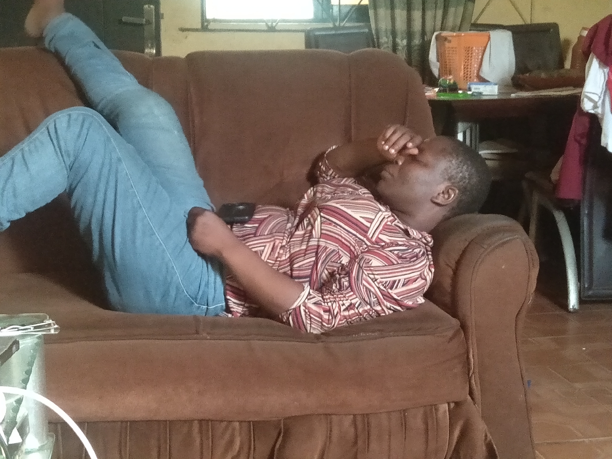 An indoor scene shows a person relaxing on a brown fabric sofa. The individual, appearing to be an adult, is lying on their back with their legs bent and raised, clad in light blue jeans. They are wearing a long-sleeved shirt with a distinctive pattern of red, white, and brown geometric stripes. One hand is placed over their eyes or forehead, suggesting they are resting, possibly fatigued, or shielding their eyes from light. A dark-colored smartphone or remote control is visible on the sofa near their midsection.
The setting appears to be a living room or a casual common area within a home in Shangisha, Nigeria. In the background, a wooden table holds an orange plastic laundry basket containing light-colored fabric, and other small items such as a white box and small green objects. A window with dark curtains is visible behind the table, providing natural light, indicating it is daytime. To the right of the frame, some clothing, including red and white garments, is draped over an unseen object. The floor has a muted, tiled or concrete appearance. The overall impression is one of a relaxed, domestic environment. No visible text is present in the image.