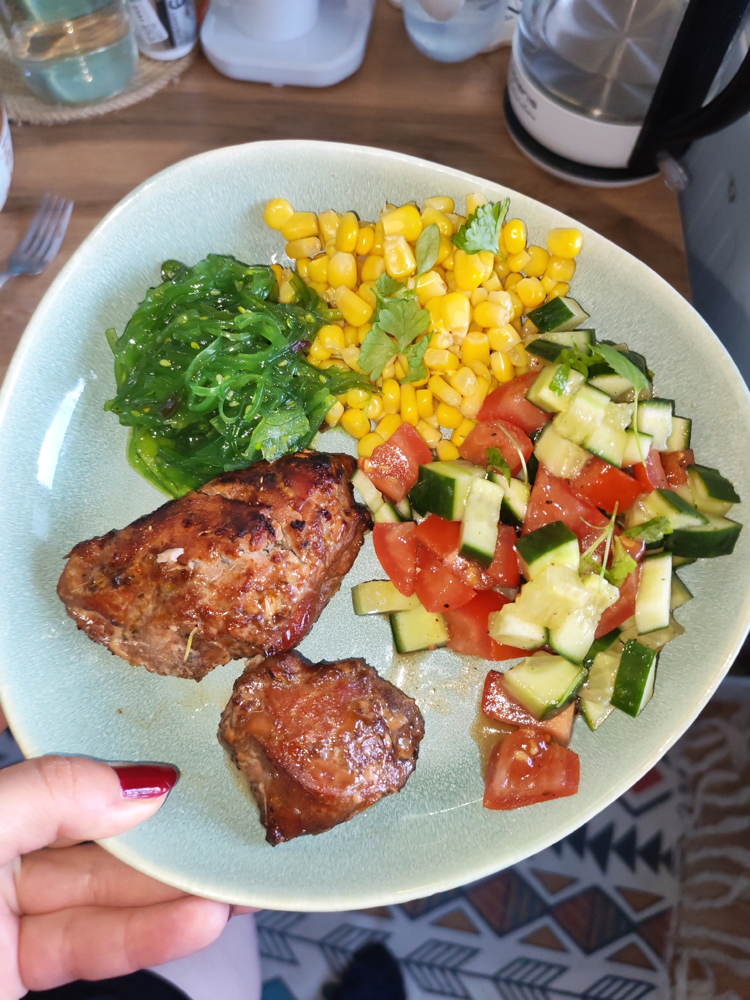 A close-up, elevated shot captures a person's hand, adorned with dark red nail polish, holding a light green plate filled with a diverse and colorful meal. The scene is set indoors, likely in a home kitchen or dining area in Izhevsk, Russia, indicated by the background elements.
On the plate, two succulent pieces of grilled, dark-brown meat, possibly pork or chicken, with a caramelized or glazed finish, occupy the lower left. Adjacent to the meat, a vibrant green portion of wakame seaweed salad, glistening with dressing and dotted with sesame seeds, is visible. Above the seaweed salad, a generous serving of bright yellow corn kernels, garnished with fresh cilantro or parsley sprigs, adds a pop of color. Completing the meal is a fresh salad composed of diced red tomatoes and green cucumbers, also lightly garnished with herbs, situated on the right side of the plate.
The background shows elements of a domestic setting: a clear glass partially filled with water on a woven coaster, a white bottle, and a silver and black electric kettle. A metal fork is also visible to the left of the plate. The overall impression is of a wholesome, freshly prepared meal. No discernible text is visible in the image.