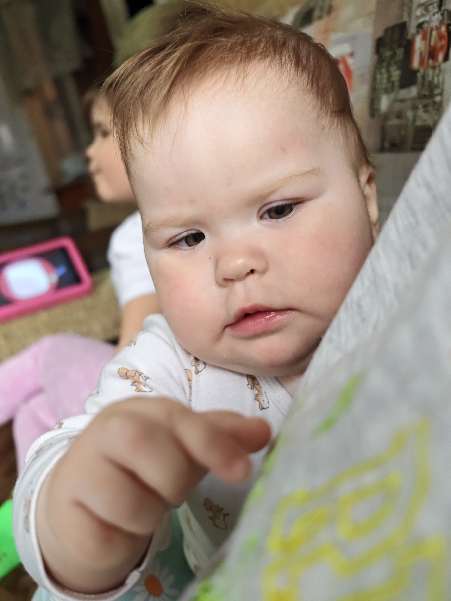 A close-up shot shows a baby with light skin, reddish-brown hair, and brown eyes looking downwards and pointing their right hand towards the camera. The baby is wearing a white onesie with a pattern of animals. Partially visible behind the baby on the left is a second child with similar features wearing a white shirt, and a pink tablet. The background includes an out-of-focus wall with what appears to be wallpaper and furniture, including what looks like a closet or cabinet, and part of a white cloth with scribbled yellow patterns. The location is unknown, but not specified as Malyi Bukor, Russia.