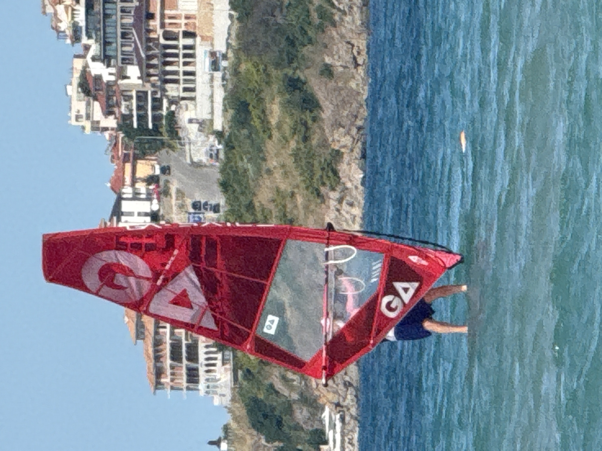 The image shows a person windsurfing in the sea, with the town of Sozopol, Bulgaria, in the background. The windsurf sail is red and has a white "GA" logo on it. The person is wearing blue shorts and is standing in shallow water. The town of Sozopol is built on a hill overlooking the sea, and many of the buildings are white with red-tiled roofs. The sky is blue and clear.