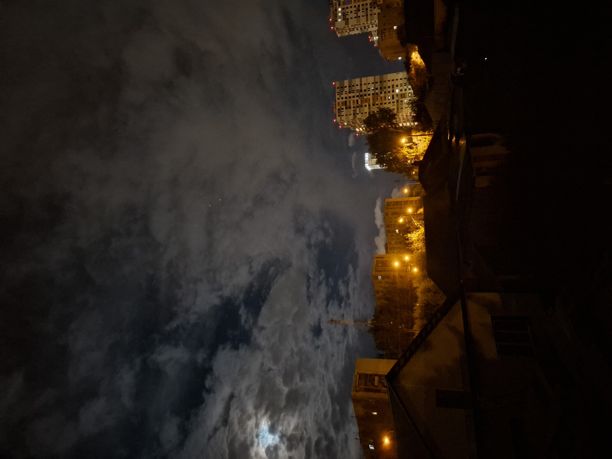 This is a nighttime photograph of a city skyline, likely in Tyumen, Russia, given the presence of Soviet-era apartment blocks. The sky is filled with dramatic, dark clouds illuminated by the moon, which is visible in the upper left corner. The clouds are dense and textured, creating a moody atmosphere. Below the sky, a cityscape unfolds. In the foreground, the dark silhouettes of smaller buildings, possibly houses or garages, are visible, their roofs sharply angled. Beyond these, larger apartment buildings with lit windows and streetlights cast a warm glow on the scene. Two tall, modern high-rises dominate the middle ground, their windows illuminated, and red lights on their rooftops indicating they are taller structures. A communications tower with an antenna is also visible. The overall impression is one of an urban environment at night, under a cloudy sky, with a mix of older and newer architecture.