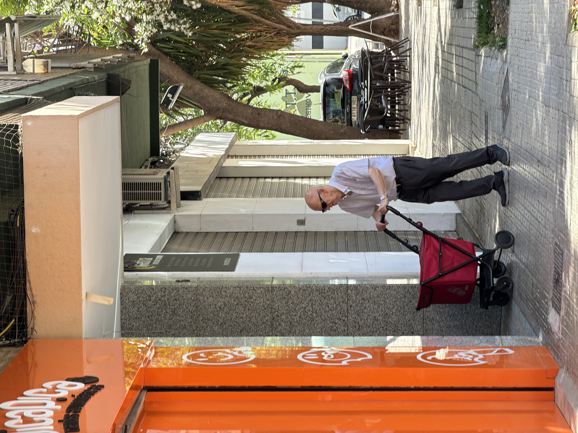 The image, rotated 90 degrees counter-clockwise, captures a sunlit street scene in Valencia, Spain. An elderly, bald man, wearing sunglasses, a light checkered short-sleeve shirt, dark trousers, and dark shoes, is walking on a paved sidewalk. He is pushing a red shopping trolley with black wheels.
The street features a row of buildings with closed roller shutters and facades in light colors, one displaying an air conditioning unit. Mature trees with green foliage line the street, casting distinct shadows that suggest bright mid-morning or late afternoon sunlight. In the background, a dark car with the license plate "6118 HBS" is parked, partially hidden by tree branches. Nearby, a stack of dark outdoor chairs indicates an establishment with an al fresco dining area. A sign behind the car shows partially visible, reversed text, possibly "YOVIGO" or "LLOYIGOV".
In the foreground, an orange, reflective street-level sign or barrier features white, stylized symbols, one resembling a face. The sidewalk is constructed from light-colored paving stones, and a grey, textured granite-like surface is visible at the base of some buildings. The scene portrays a calm, routine moment in an urban setting.