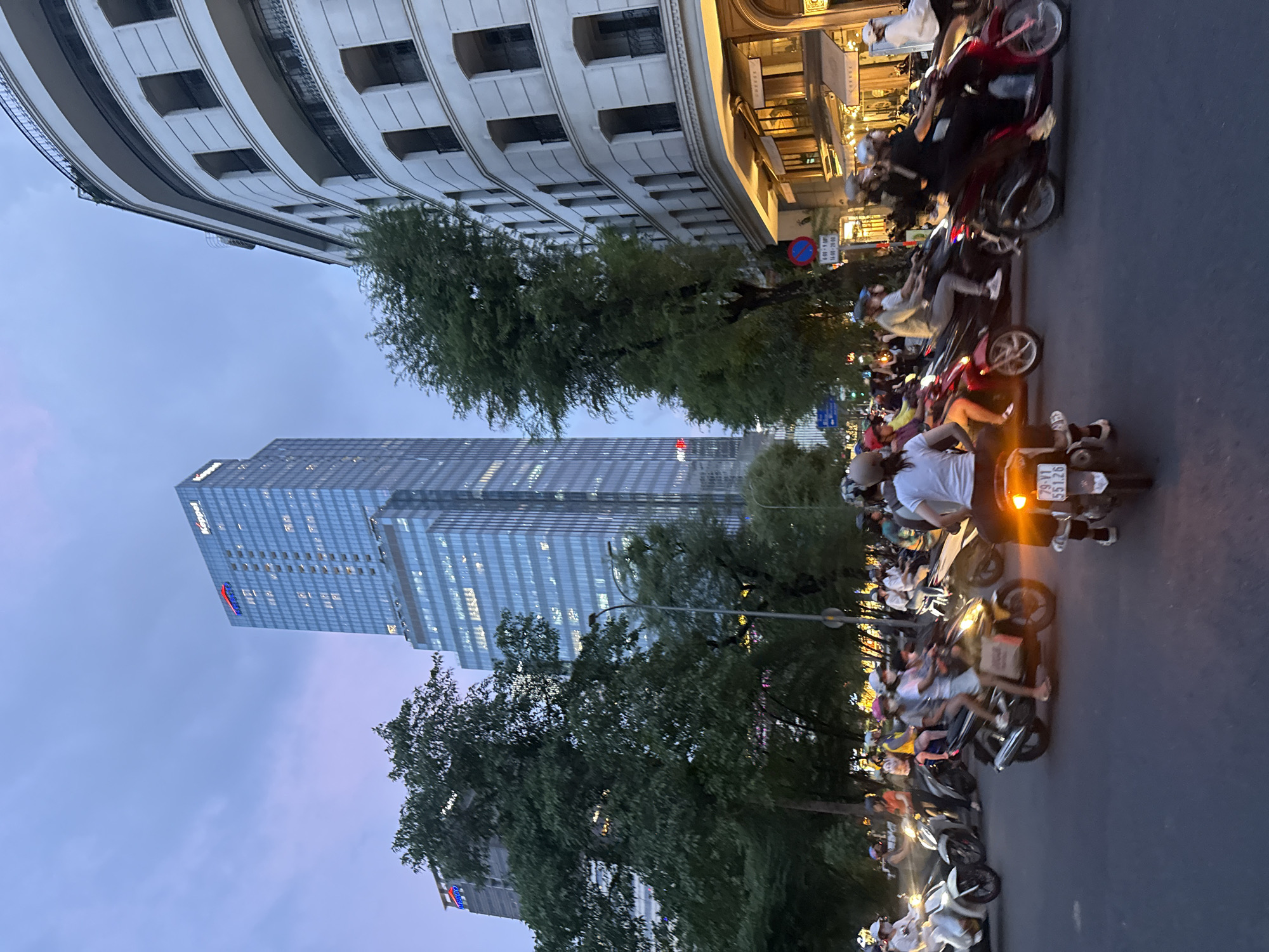 The image captures a street scene in the city of Thủ Đức, Vietnam, at dusk. A multi-lane road is filled with a considerable number of motorbikes, many carrying two or more passengers. Most individuals are wearing helmets, and some are wearing masks. The traffic appears to be moving, with some motorbikes illuminated by their headlights. In the background, a modern, tall skyscraper with numerous windows dominates the skyline, displaying the "KISSAGO" and "Kippel" logos. Adjacent to the skyscraper is a white, multi-story building with curved facades and balconies. Trees line the street, and streetlights are visible, indicating the onset of evening. A traffic sign is also present on a pole. The overall atmosphere suggests a busy urban environment during a transitional period of the day.
