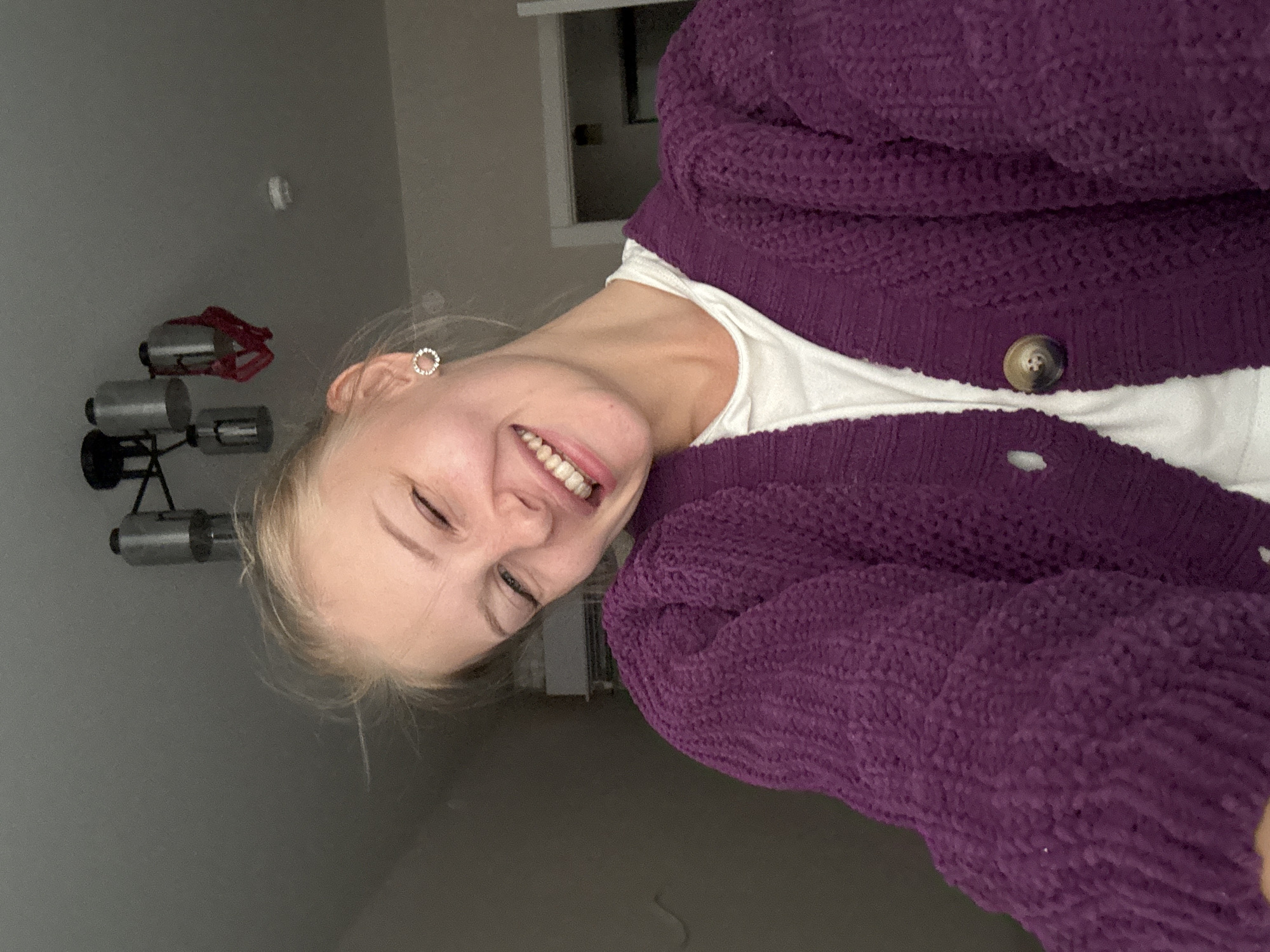 The image shows a young Caucasian woman with blonde hair and fair skin, winking and smiling. She is wearing a purple knitted cardigan over a white t-shirt and small circular earrings. The background is a room with a grey ceiling, a chandelier, and a door. The image is a selfie taken indoors. It is not possible to determine the location in the city of Perm, Russia, from the provided image.