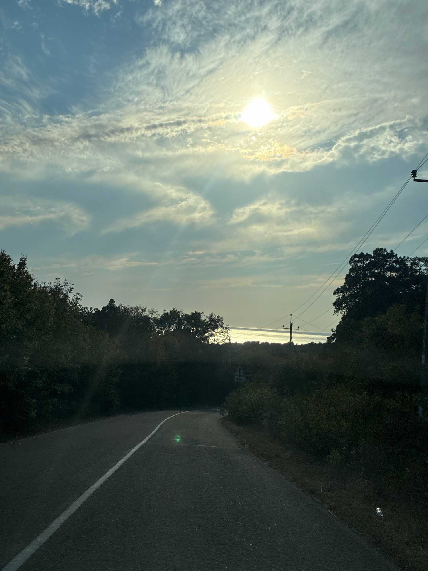 A low-angle, wide shot captures a paved road with a white line marking its center, curving to the left through a landscape of trees and vegetation. The road surface appears slightly wet. In the background, a body of water, possibly a sea or large lake, reflects the sunlight. The sky is a pale blue, filled with wispy clouds and a bright sun partially obscured by them, casting rays of light. Power lines and utility poles are visible along the right side of the frame, extending towards the horizon. The environment suggests a rural or semi-rural setting, with dense foliage lining the road and a distant body of water. There are no people visible in the image. A triangular sign with the number "50" and "km/h" is partially visible on the left side of the road.