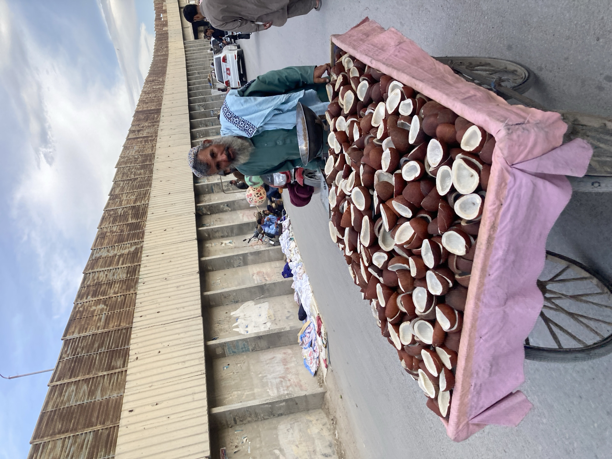 The image shows a man in Quetta City Tehsil, Pakistan, standing next to a cart overflowing with halved coconuts, likely for sale. He has a gray beard and wears a traditional cap and clothing. The cart is covered with a pink cloth. Behind him, there is a corrugated metal structure, what appears to be stairs, and other people walking on the street. A white car is also visible in the background. The scene depicts street vending and everyday life.