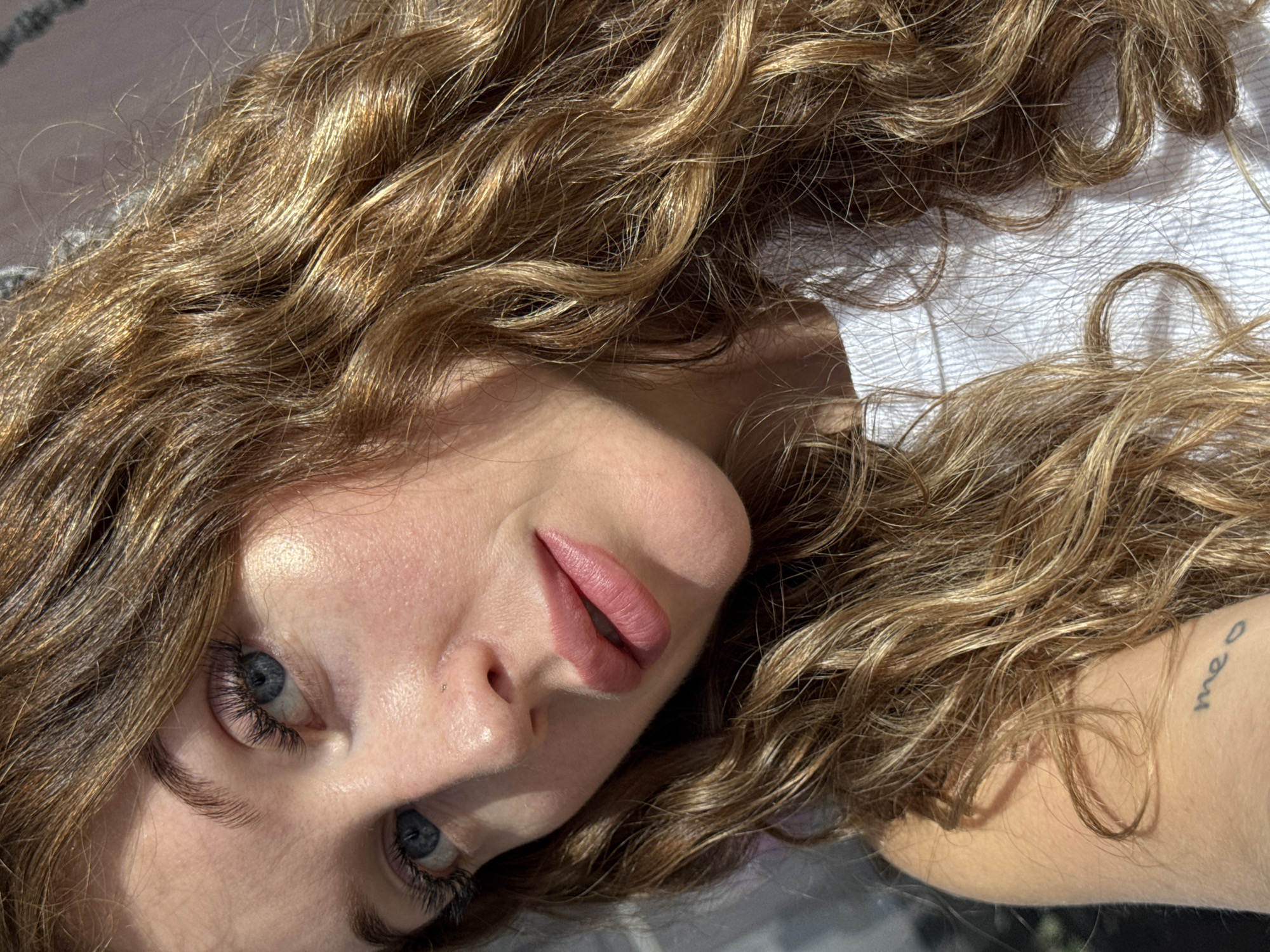 A close-up portrait of a young woman with fair skin, blue eyes, and wavy, light brown hair is presented. She appears to be lying down, facing the camera at an angle. Her lips are pink and full, and her eyelashes are long and dark. Part of a white garment is visible behind her hair. A small tattoo is visible on her inner wrist. The image exhibits a shallow depth of field, focusing primarily on her face. The location is not discernible from the image.