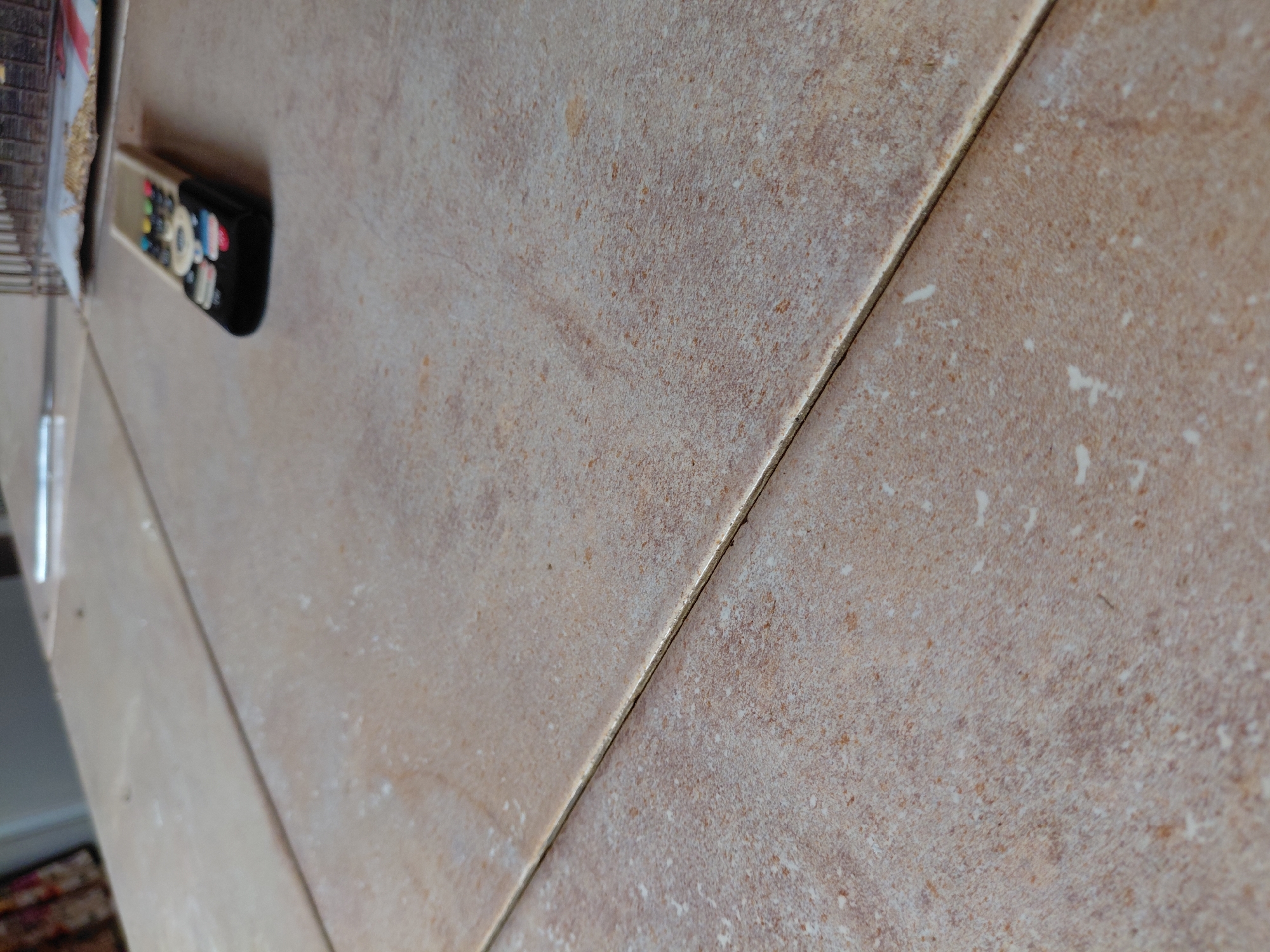 The image captures a close-up, angled view of a tiled surface, likely a floor, in what appears to be an indoor setting. The tiles are rectangular, in shades of light brown and beige, with a mottled, slightly textured pattern that includes darker brown specks and lighter, off-white patches. A narrow grout line, lighter than the tiles, separates the individual pieces.
In the upper left portion of the image, resting on one of the tiles, is a remote control. It is black with numerous small, multi-colored buttons, primarily white, black, red, blue, and yellow. The remote control is somewhat out of focus compared to the foreground tiles and grout lines.
To the far left, partially obscured and out of focus, is a vertical silver-colored element, possibly a leg of furniture or a stand, and a glimpse of what might be books or other items on a shelf. The lighting is soft and ambient, casting minimal shadows. No human figures or discernible activity are present. No legible text is visible in the frame. The overall impression is one of a domestic or interior space.