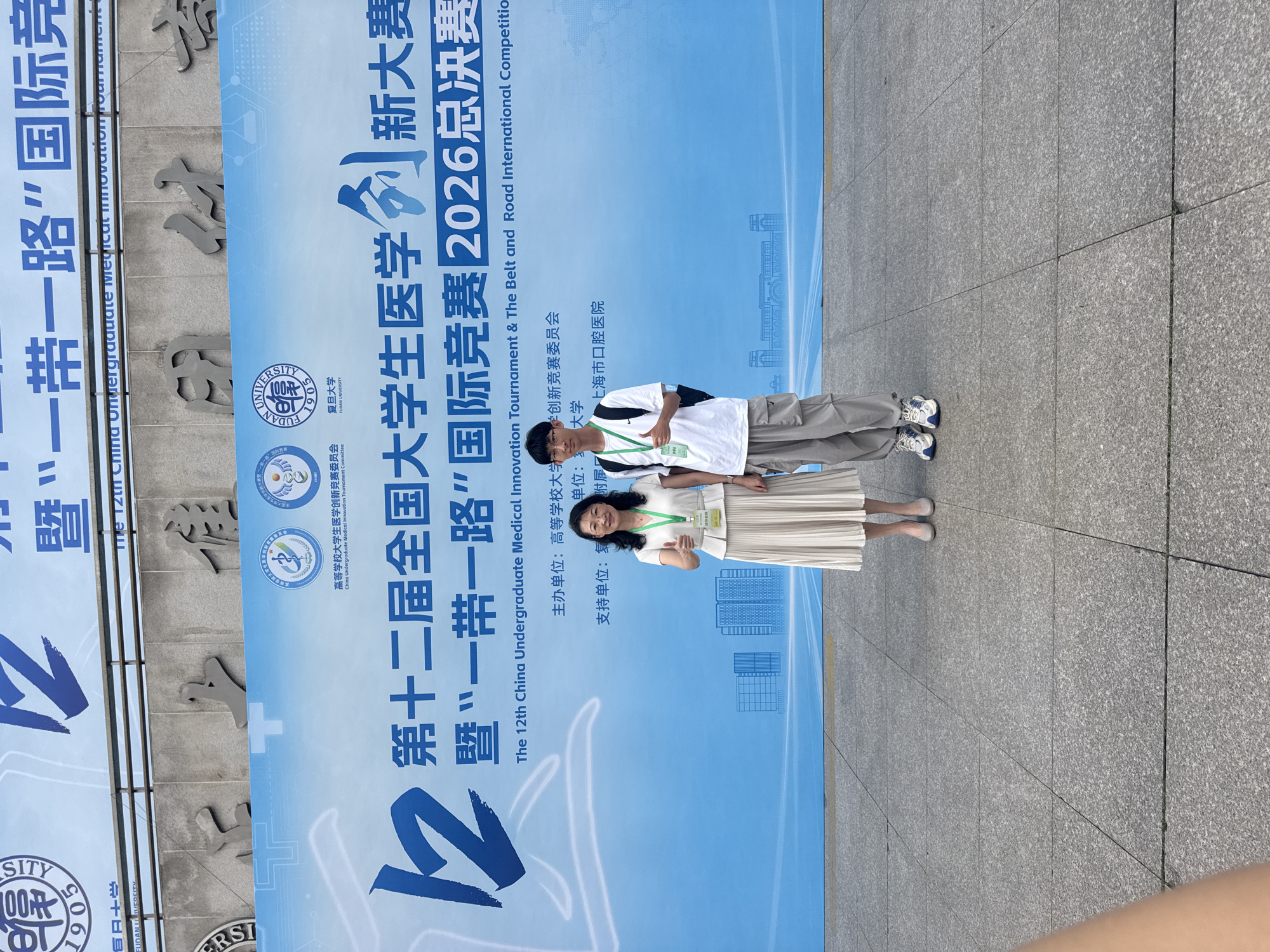Two individuals, an adult female and a young male, stand in front of a large blue banner. The banner features Chinese text and English text that reads "The 12th China Undergraduate Medical Innovation Tournament & The Belt and Road International Competition 2026 Finals". The female, on the left, is wearing a white dress and white heels, with a lanyard around her neck. She is giving a thumbs-up gesture. The male, on the right, is wearing a white t-shirt, grey cargo pants, and white sneakers. He is also wearing a lanyard and giving a thumbs-up. The background banner has logos of universities. The location appears to be an outdoor urban setting, possibly a campus or event venue in Yangpu District, China, indicated by the university logos and the banner's content. The ground is paved with granite tiles.