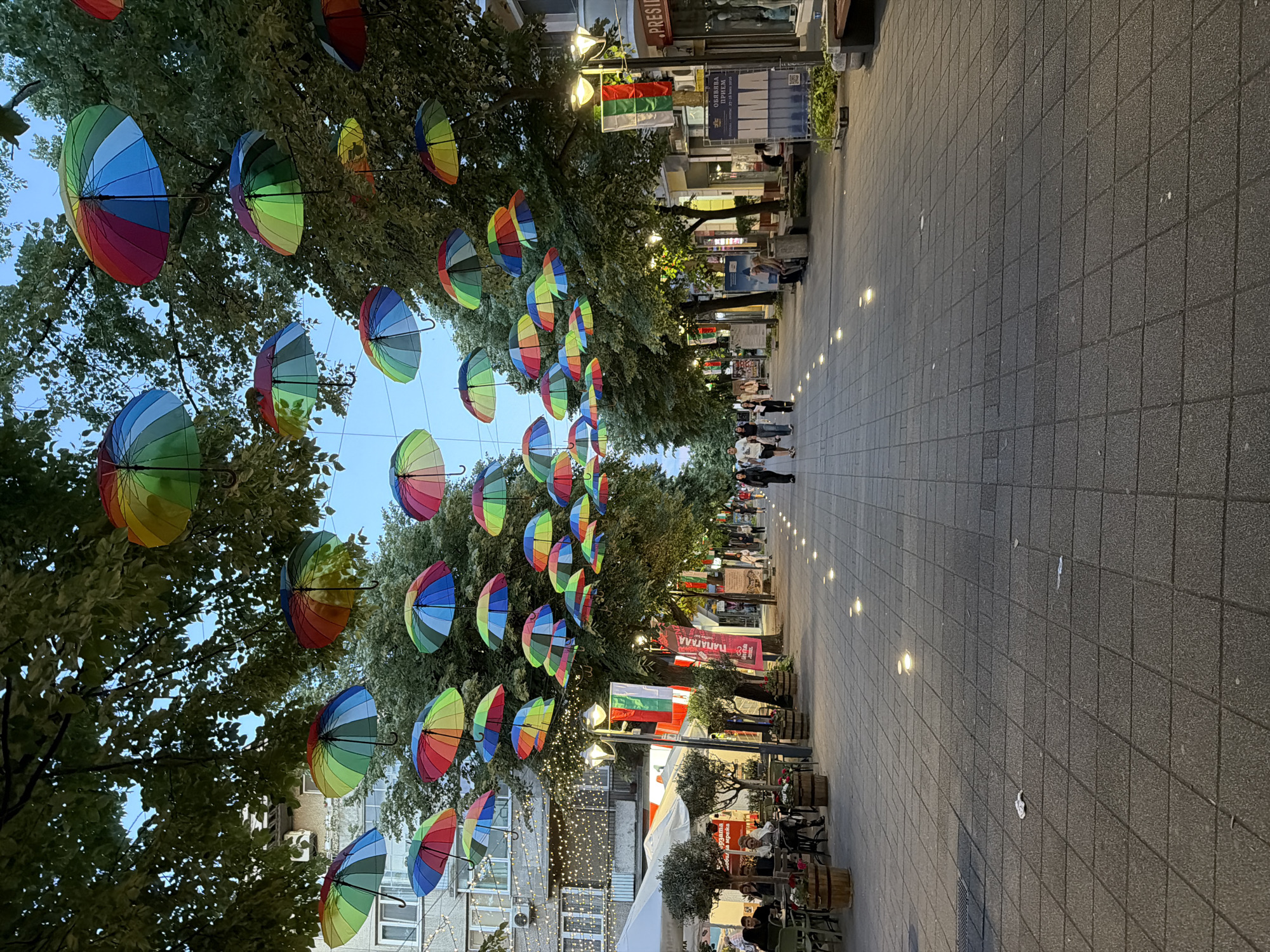 The image shows a pedestrian street in Burgas, Bulgaria, identifiable by the Bulgarian flag hanging prominently along the street. Above the street, rows of colorful, striped umbrellas are suspended, creating a canopy effect between trees. People can be seen walking down the street. The walkway is paved with dark, rectangular tiles. Small lights are embedded in the pavement, running along the center of the path. Buildings and storefronts line both sides of the street. Outdoor seating areas are visible near the buildings, with some tables occupied. The sky is visible between the trees and umbrellas.