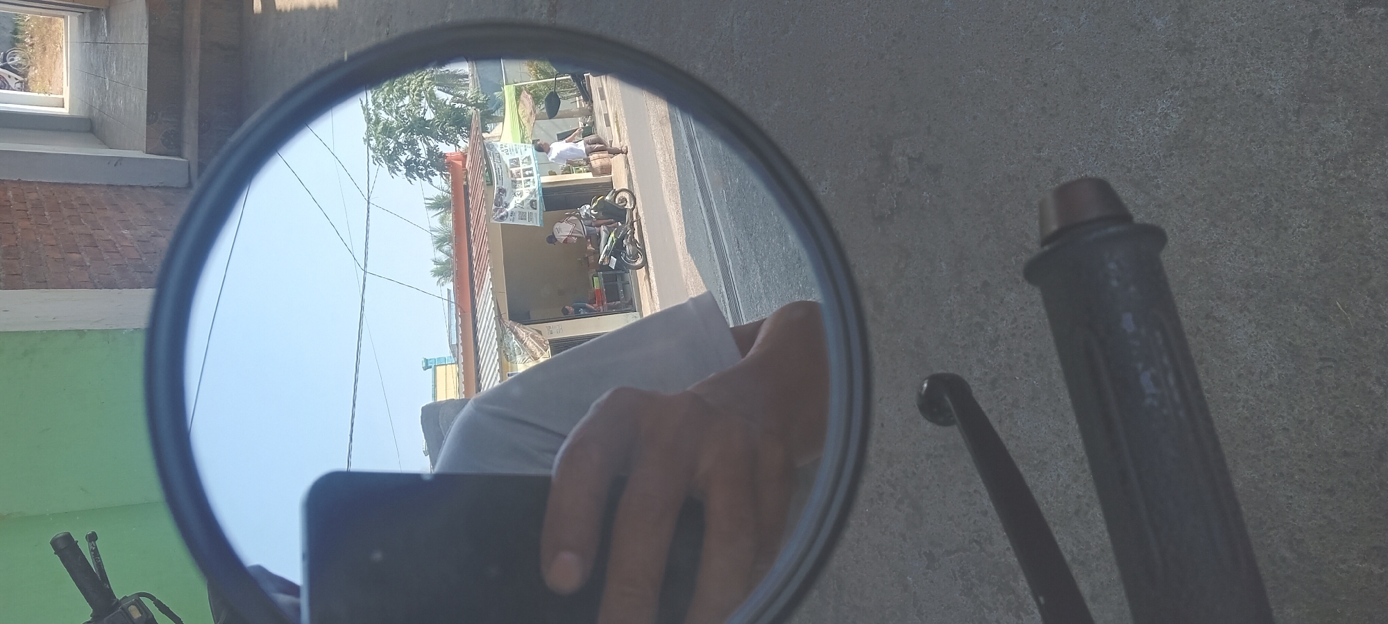 The image is a reflection in a motorcycle's rearview mirror, taken in Tempurejo, Indonesia. The foreground shows parts of a motorcycle, including a handlebar grip. The reflected scene shows a street with a building on the right and a row of shops on the left. Several people are visible in the reflection. One person is walking on the sidewalk. Two men are standing near a green motorcycle parked in front of a shop. Another person, possibly a child, is visible inside the shop. The sky is clear and blue. The building on the right appears to be a house with a brick wall and a doorway. The shops on the left have corrugated metal roofs and are decorated with banners. The overall impression is a snapshot of daily life on a street in a small Indonesian town.