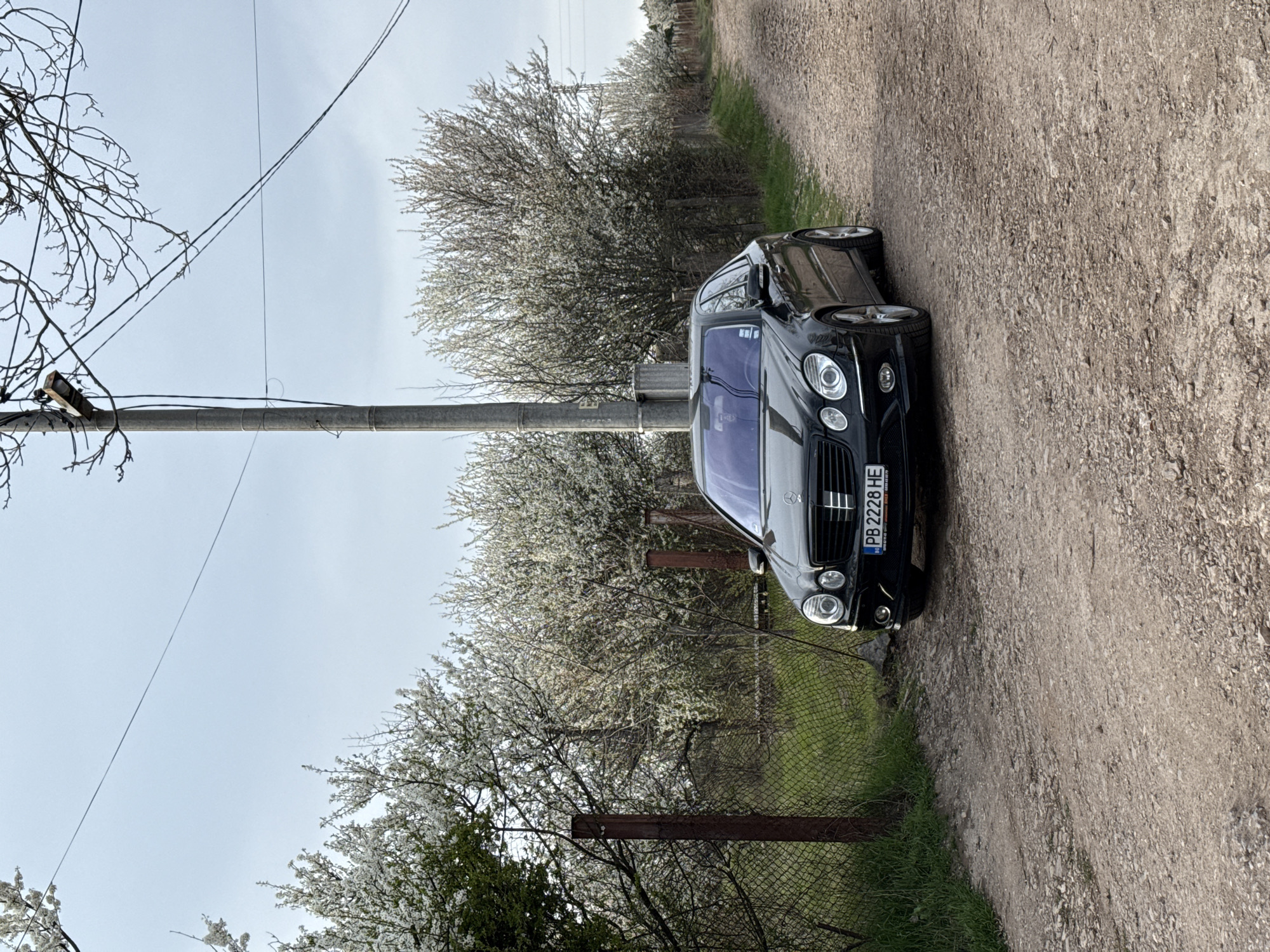 A black Mercedes-Benz E-Class is parked on a dirt road next to a wire fence and blossoming trees in Buhovo, Bulgaria. A concrete utility pole stands to the left of the car, with power lines overhead. The car's license plate reads "PB 2228 HE". The surrounding vegetation includes grass and trees with white blossoms. The sky is overcast.
