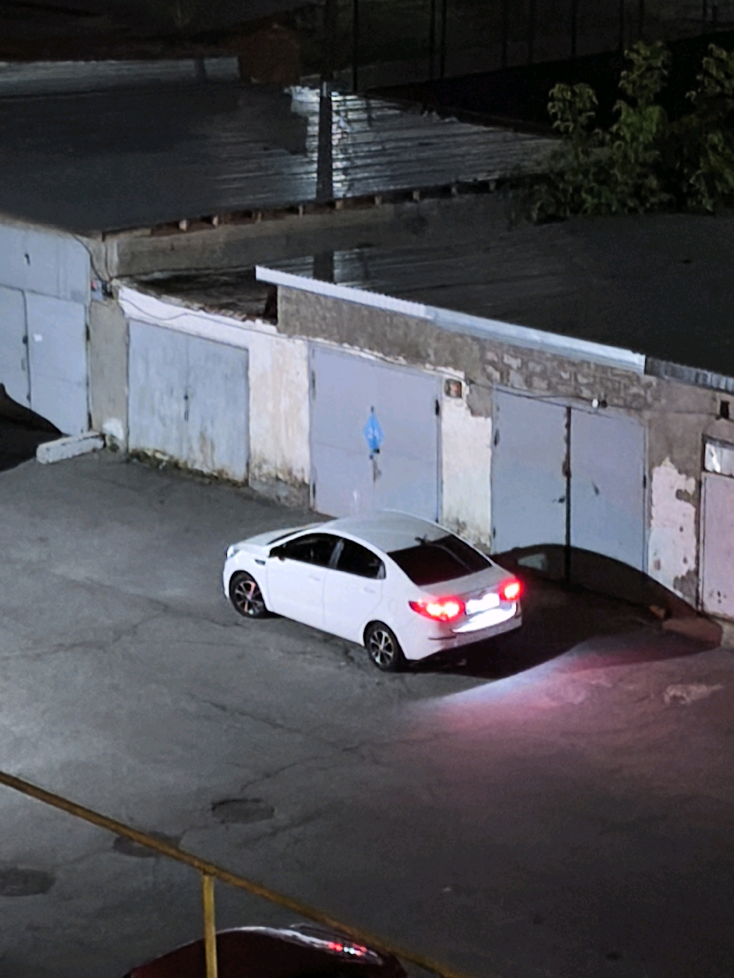 A white sedan is parked in front of a row of garages at night. The car's taillights are illuminated, casting a pinkish glow on the asphalt. The garages are made of concrete and metal, and some appear to be old and weathered. There are trees growing behind the garages. The scene is likely located in a residential or industrial area.