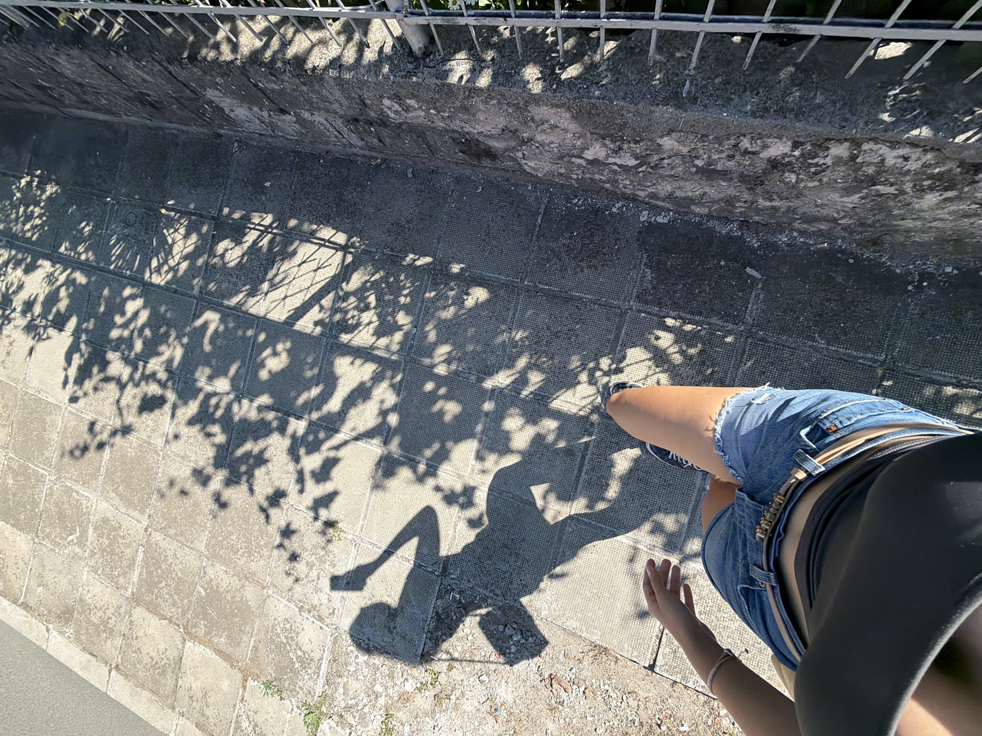 A first-person perspective shot shows a person wearing denim shorts and a dark top walking on a tiled sidewalk. The person is holding a smartphone in their right hand, creating a shadow on the ground that depicts them taking a selfie. The sidewalk is bordered by a rough concrete wall on the right, with a metal fence above it. Tree shadows are cast across the sidewalk. The location is the city of Tsarevo, Bulgaria.