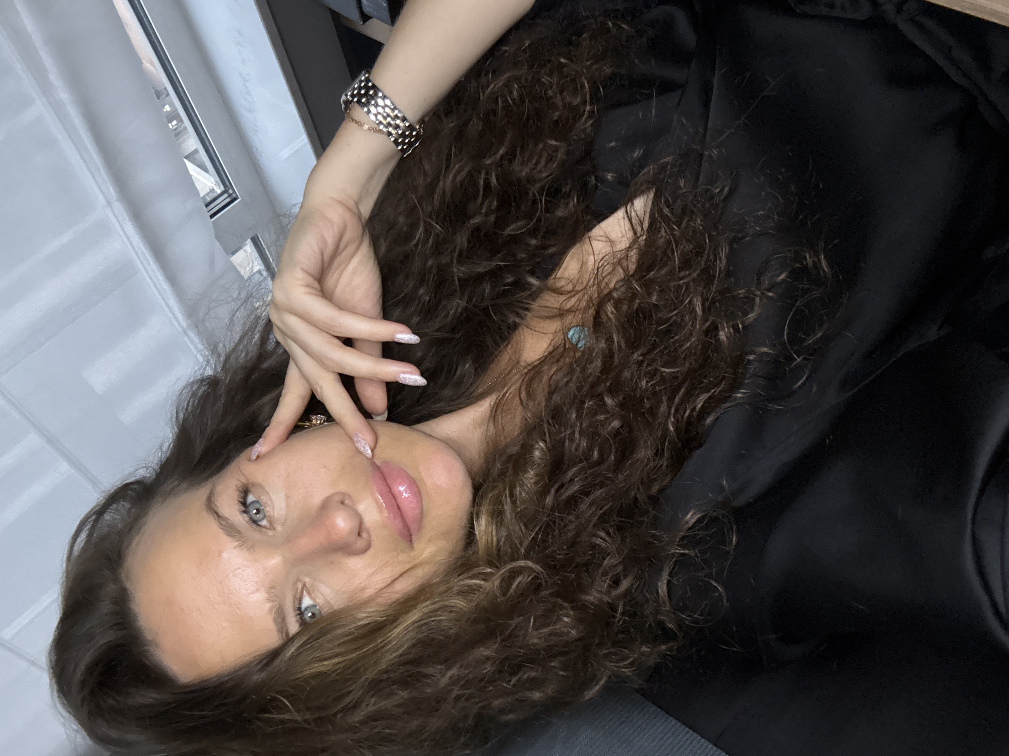 The image shows a medium shot of a woman with long, wavy brown hair. She is wearing a black garment that appears to be a robe or dress. Her left hand is resting on her cheek, and her right arm is bent, with her hand resting near her head. She is wearing a silver-toned wristwatch on her right wrist. The background is out of focus but appears to be an indoor setting with light-colored walls or blinds and a window. There is no specific indication of the location being Moscow, Russia. The woman's makeup is subtle, with emphasis on her lips and eyes. The overall impression is of a casual, personal photograph.