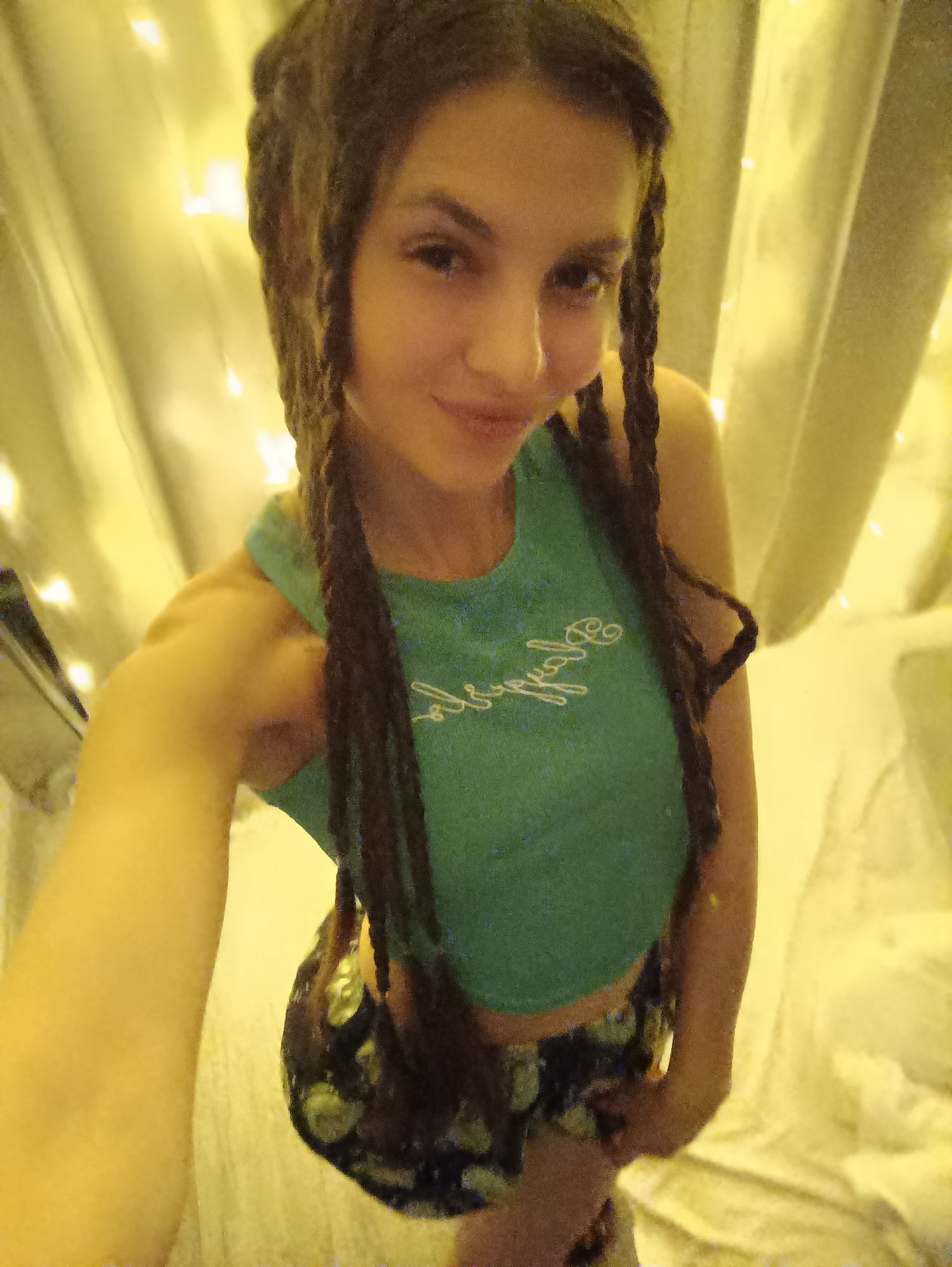 A selfie of a woman with brown braided hair, wearing a green tank top with white lettering and patterned shorts. The background features white curtains illuminated by yellow fairy lights. Location: Unknown, Unknown.