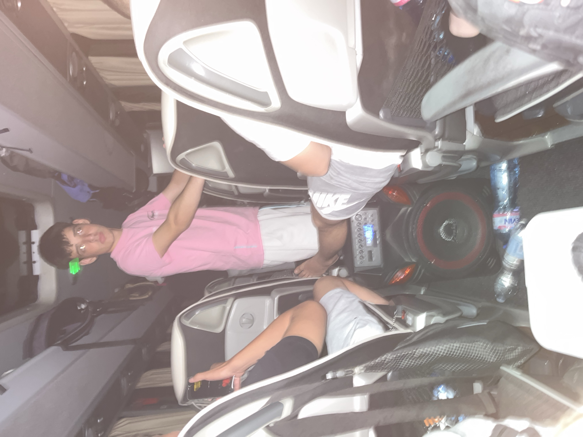 The image depicts the interior of a vehicle, possibly a bus or van, with passengers seated in reclining chairs. There are two individuals visible; one wearing a pink shirt and grey Nike shorts, and another wearing a grey shirt and black shorts. A water bottle and a speaker are also visible. The exact location is not discernible from the image itself. There is no information available to ascertain that the location is Vinogradets, Bulgaria.