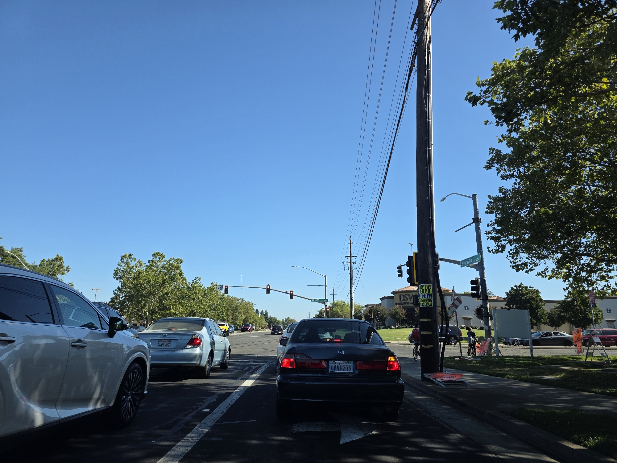This image captures a street scene in Elk Grove, USA, on a clear, sunny day, likely in the afternoon due to the bright sunlight and long shadows. The setting is a multi-lane road with moderate traffic.
The primary subjects are vehicles stopped in traffic. In the foreground, a white SUV is partially visible on the left. Behind it, a black sedan with a California license plate reading "LAVH279" is prominently featured. Further down the road, several other cars are visible, including a light blue sedan with a license plate that appears to read "5LBY000."
On the right side of the road, a sidewalk is present, with a power pole carrying multiple wires. A street lamp is also attached to the pole. A person is seen cycling on the sidewalk, and another person appears to be standing near the road. In the background, buildings with signage are visible, including one that reads "DSW." Trees line the side of the road, providing shade. Traffic lights are visible at an intersection ahead, indicating that the vehicles are stopped at a red light.
Notable details include the clear blue sky, the bright sunshine casting shadows, and the presence of traffic. The overall atmosphere appears to be one of a typical, busy day on a suburban or commercial street. The visible text includes the license plates of the cars, street signs (one appearing to say "Bruceville Rd"), and some flyers or signs attached to the power pole.