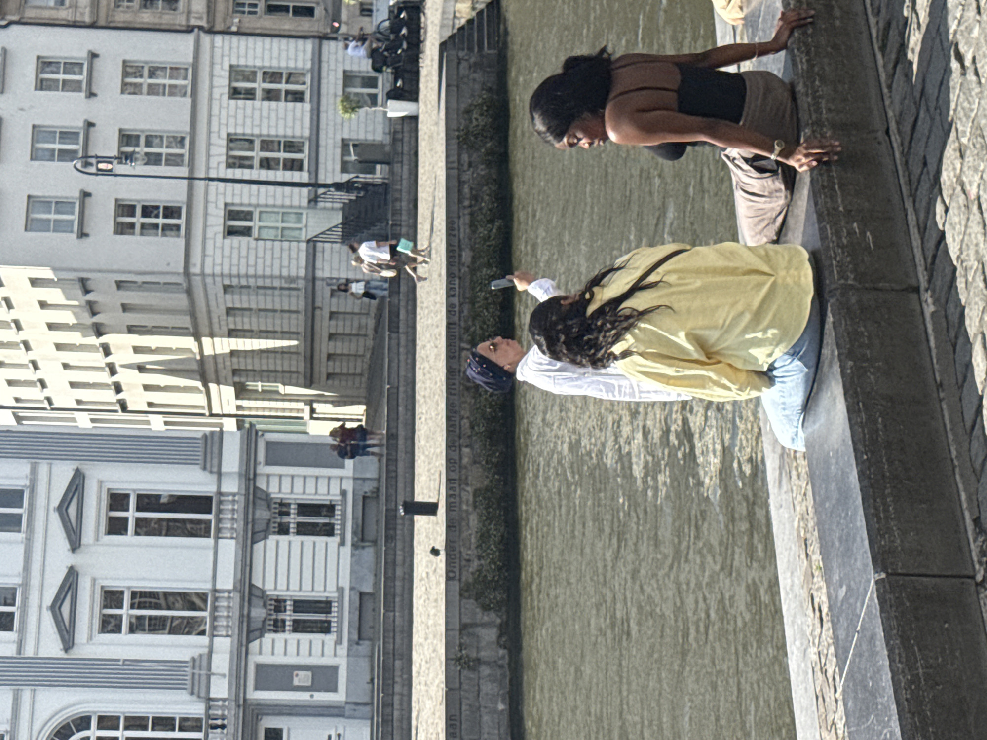 The image depicts a scene along a canal in Ghent, Belgium, likely near the Graslei or Korenlei, given the presence of historic architecture. In the foreground, three young women are seated on a stone embankment overlooking the water. One woman, on the left, has long dark curly hair and is wearing a yellow jacket over blue jeans. She is facing away from the camera. Next to her, on the right, is another young woman with dark hair, wearing a black tank top and a tan skirt, and is looking to her left. In the background, a third woman, wearing a white shirt and a dark headscarf, is holding up a smartphone, possibly taking a photo or video of the other two women. The canal is calm with ripples on its surface. Behind the embankment, a paved walkway is visible, with several people walking in the distance. The architecture in the background consists of multi-story buildings with large windows and classical detailing, typical of Ghent's historic city center. The lighting suggests it is a bright, sunny day.