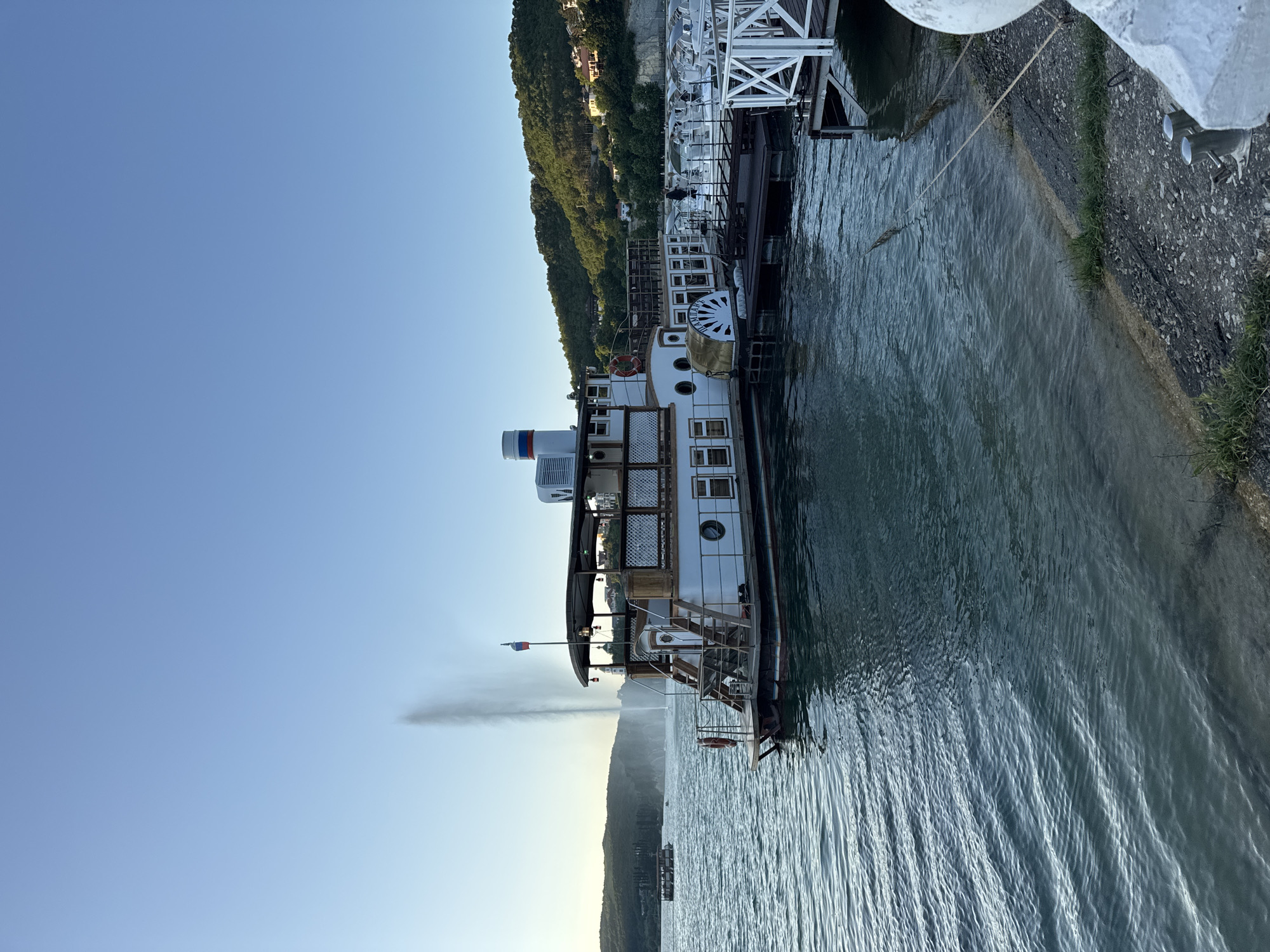 The image shows a paddlewheel steamboat docked at a pier on a body of water, likely Abrau Lake in Abrau-Dyurso, Russia. The sky is clear and blue, indicating it is daytime, possibly late afternoon given the warm lighting on the hills. The steamboat is white with wooden accents and features a covered deck, stairs, and a paddlewheel on its side. A Russian flag is visible on the stern. In the background, a tall water fountain sprays water into the air, and a lush, green hillside with scattered buildings rises behind the lake. Other boats and a pier structure are also visible to the right of the steamboat. The water is calm with gentle ripples. The foreground shows a shoreline with gravel and some grass.